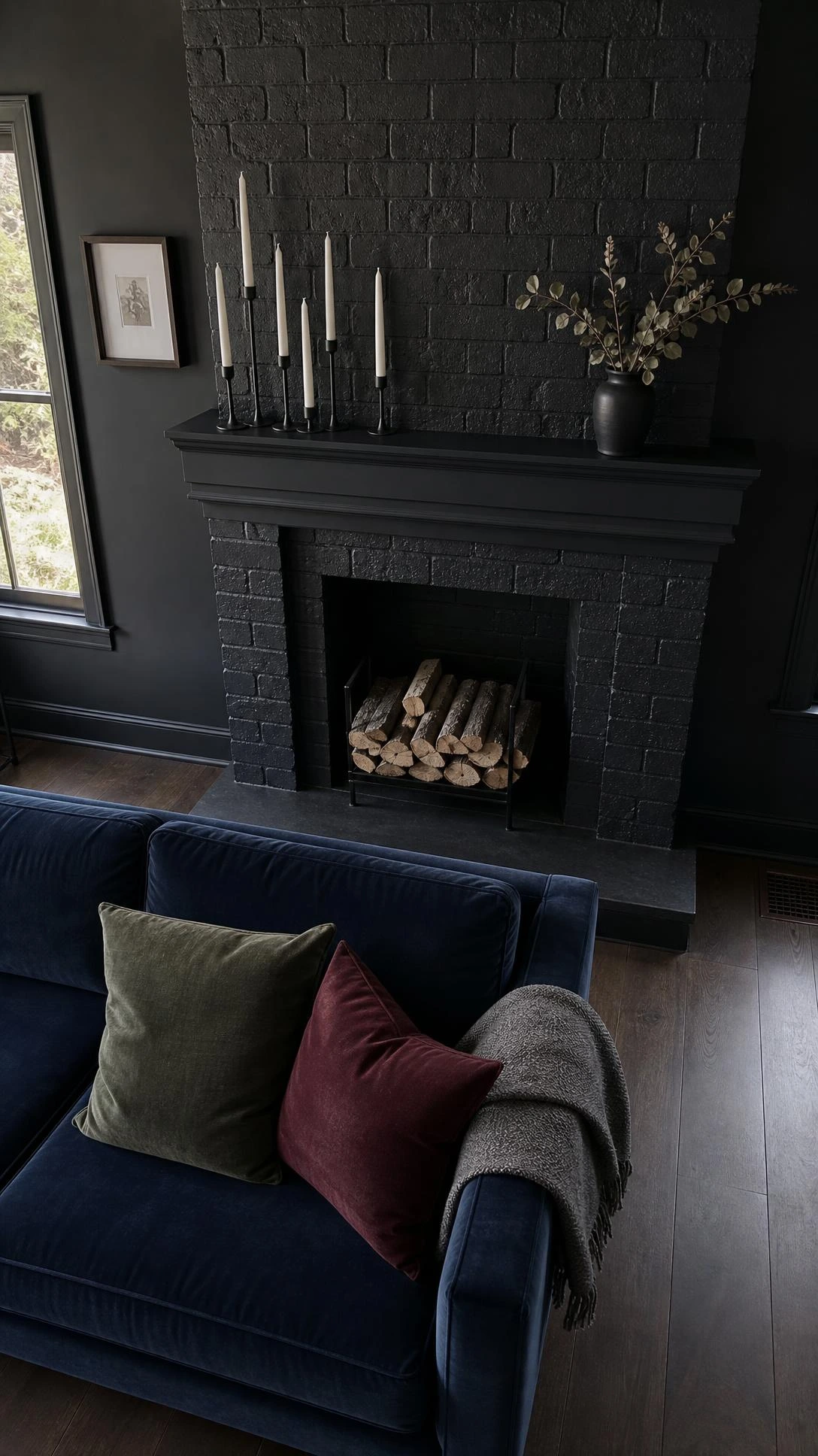 Deep charcoal-painted brick fireplace with visible rough texture anchoring a moody living room, natural overcast light highlighting the dark matte finish