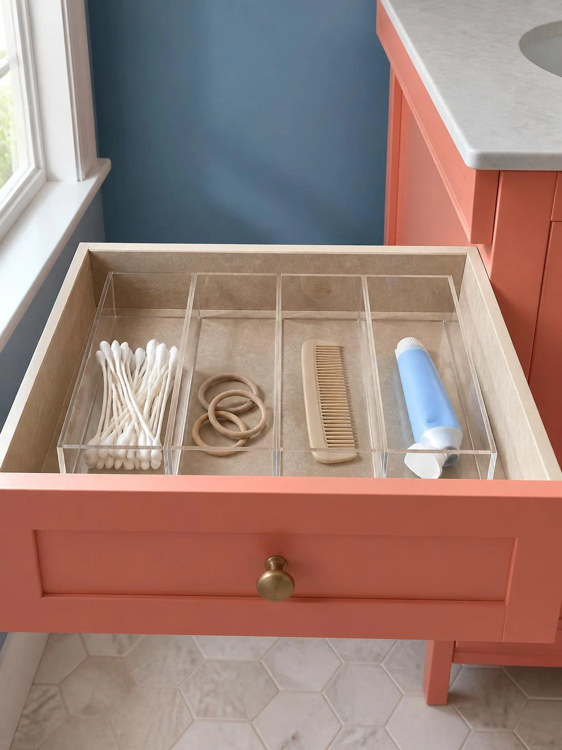 Clear acrylic drawer organizer inserts dividing a coral vanity drawer into tidy compartments holding cotton swabs, hair ties, a comb, and toothpaste