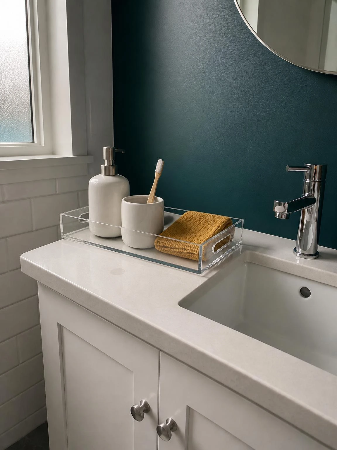 Clear acrylic tray on a teal-walled bathroom vanity holding a white soap pump, ceramic toothbrush cup, and folded mustard face cloth — a budget small bathroom organization hack