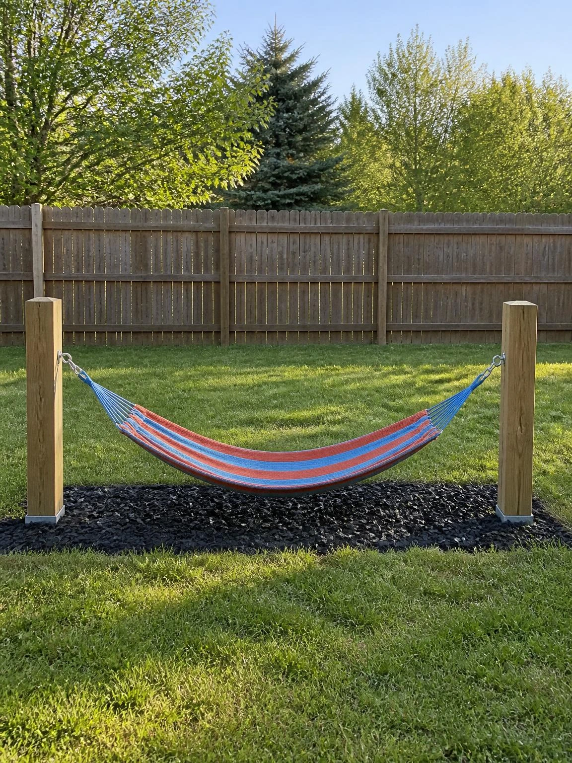 Cotton-weave hammock in rust and blue stripes hung low between timber posts over rubber mulch, creating a safe backyard hammock lounge scaled perfectly for kids