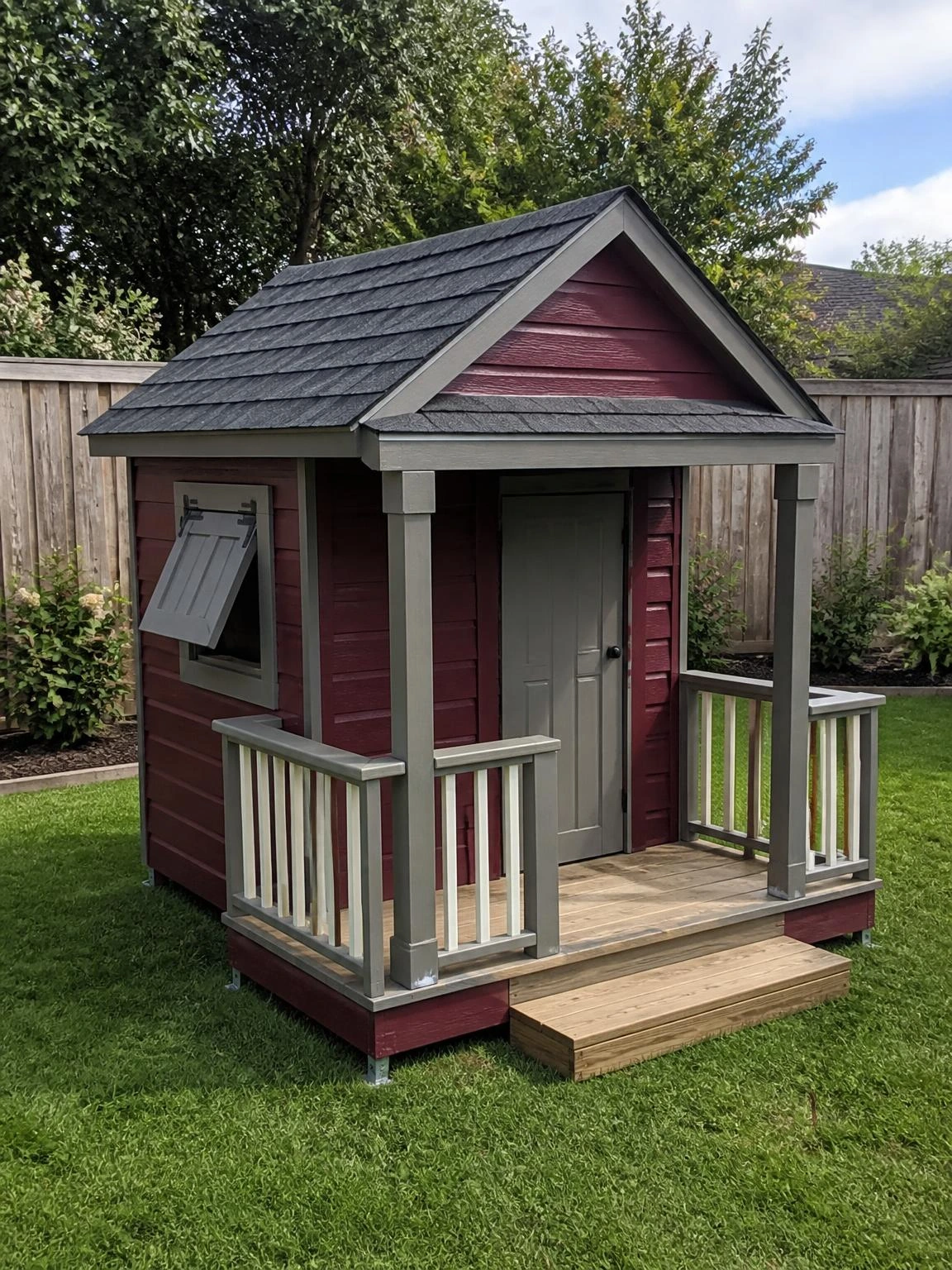 Burgundy timber backyard playhouse with gray trim, covered porch, and charcoal shingle roof standing on a trimmed suburban lawn