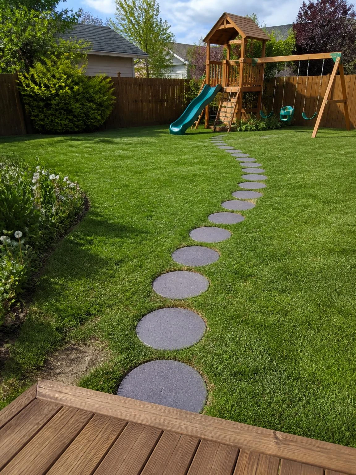 Plum-gray concrete stepping stones curve across a lush backyard lawn, spaced for kids' strides and leading from a timber patio to a cedar play structure