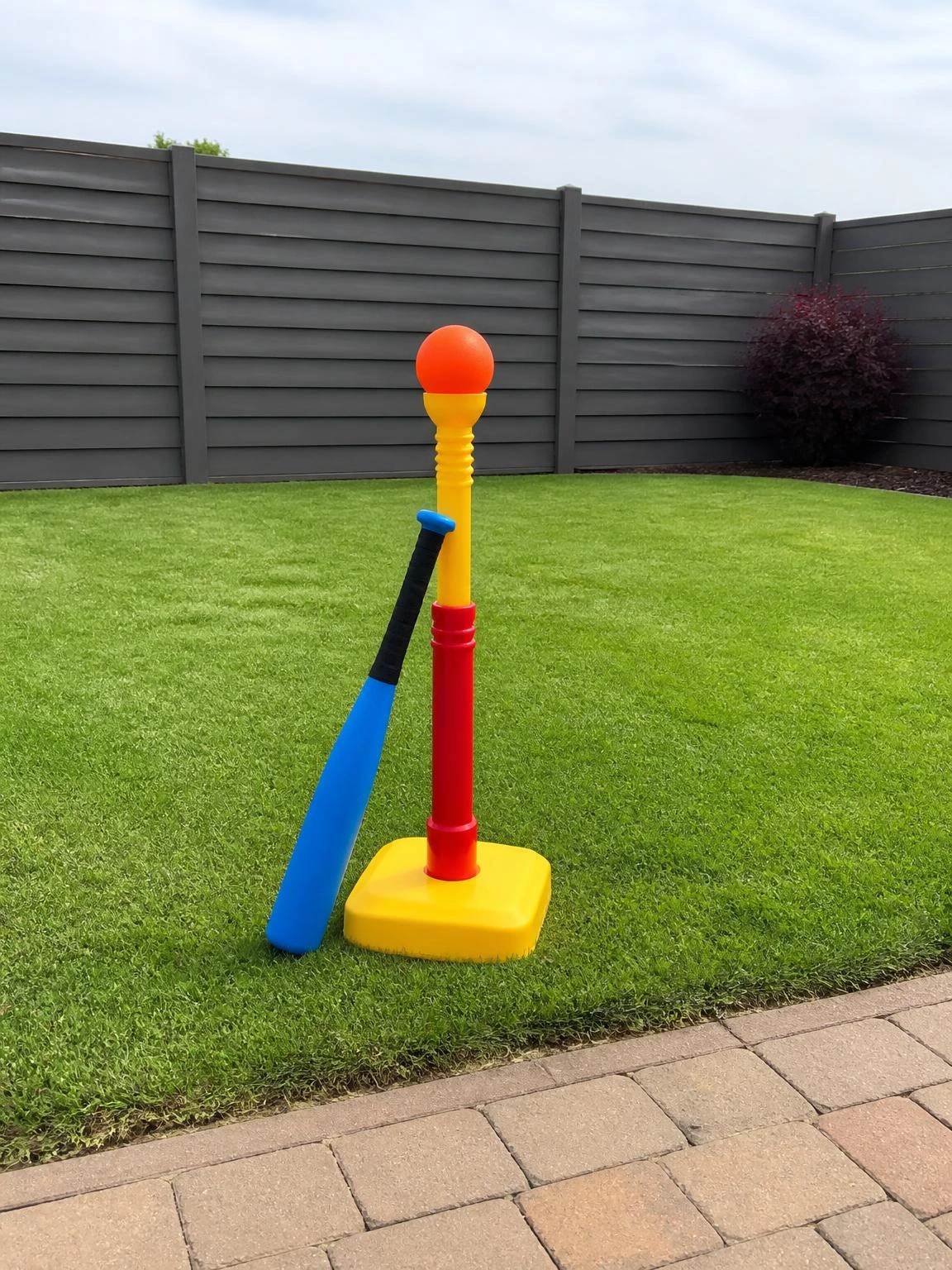 Bright yellow and red adjustable tee-ball stand with foam bat and soft rubber ball on a neatly mown backyard lawn, ready for kids to practice hitting