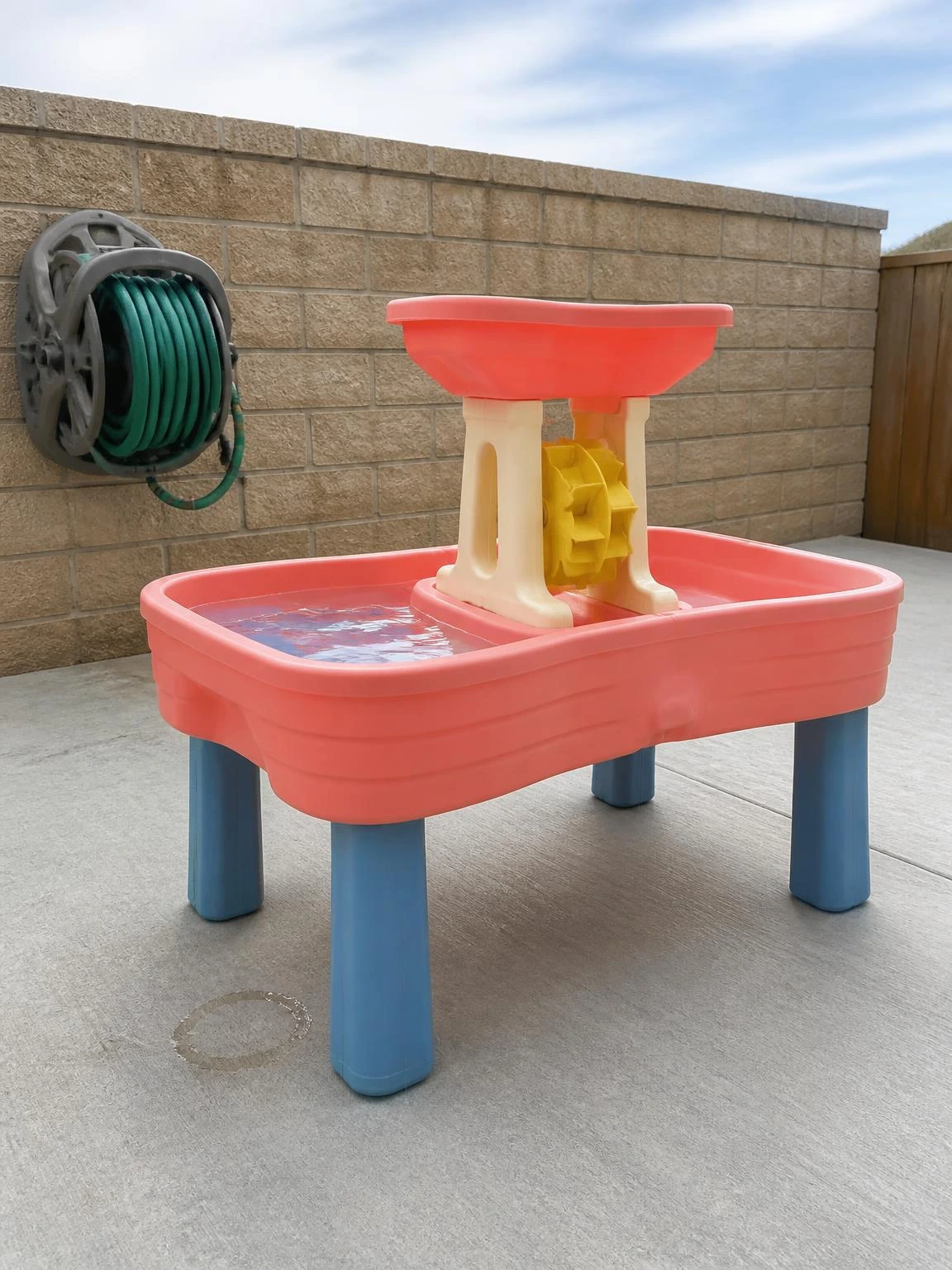 Coral plastic water table with dual basins and yellow waterwheel on a sealed concrete patio, a simple backyard water table setup for toddlers