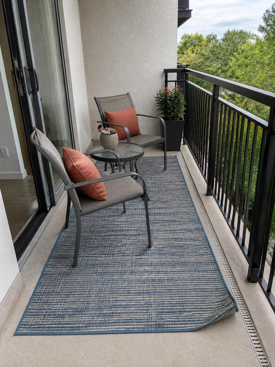 Measured balcony outdoor rug zone with a blue-and-stone rug, metal chairs, glass table, and clear drainage margins
