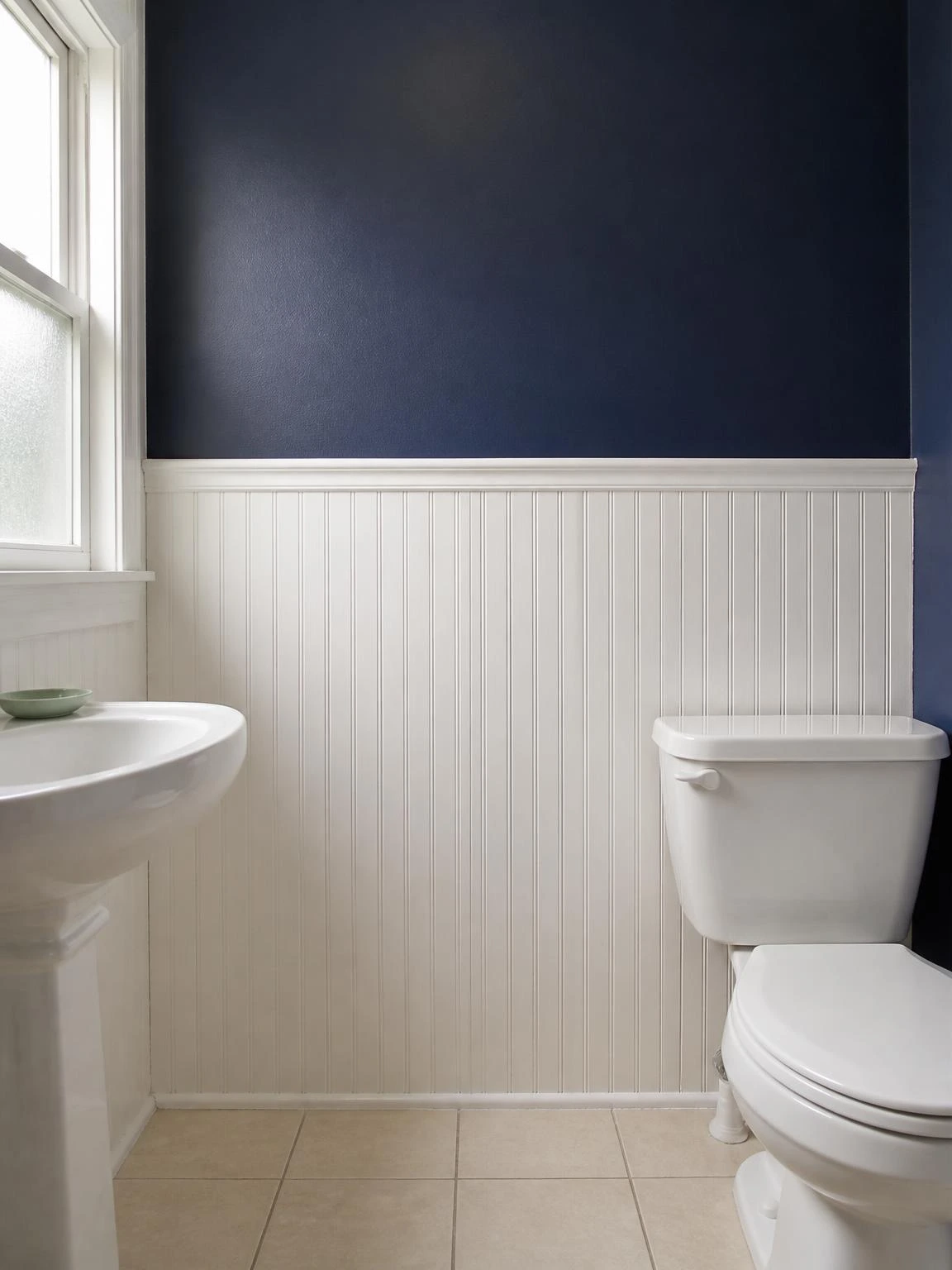 White-painted MDF beadboard panels cover the lower wall of a small bathroom, topped by a chair-rail cap strip against deep navy upper walls — a budget wainscoting idea that adds structure and style