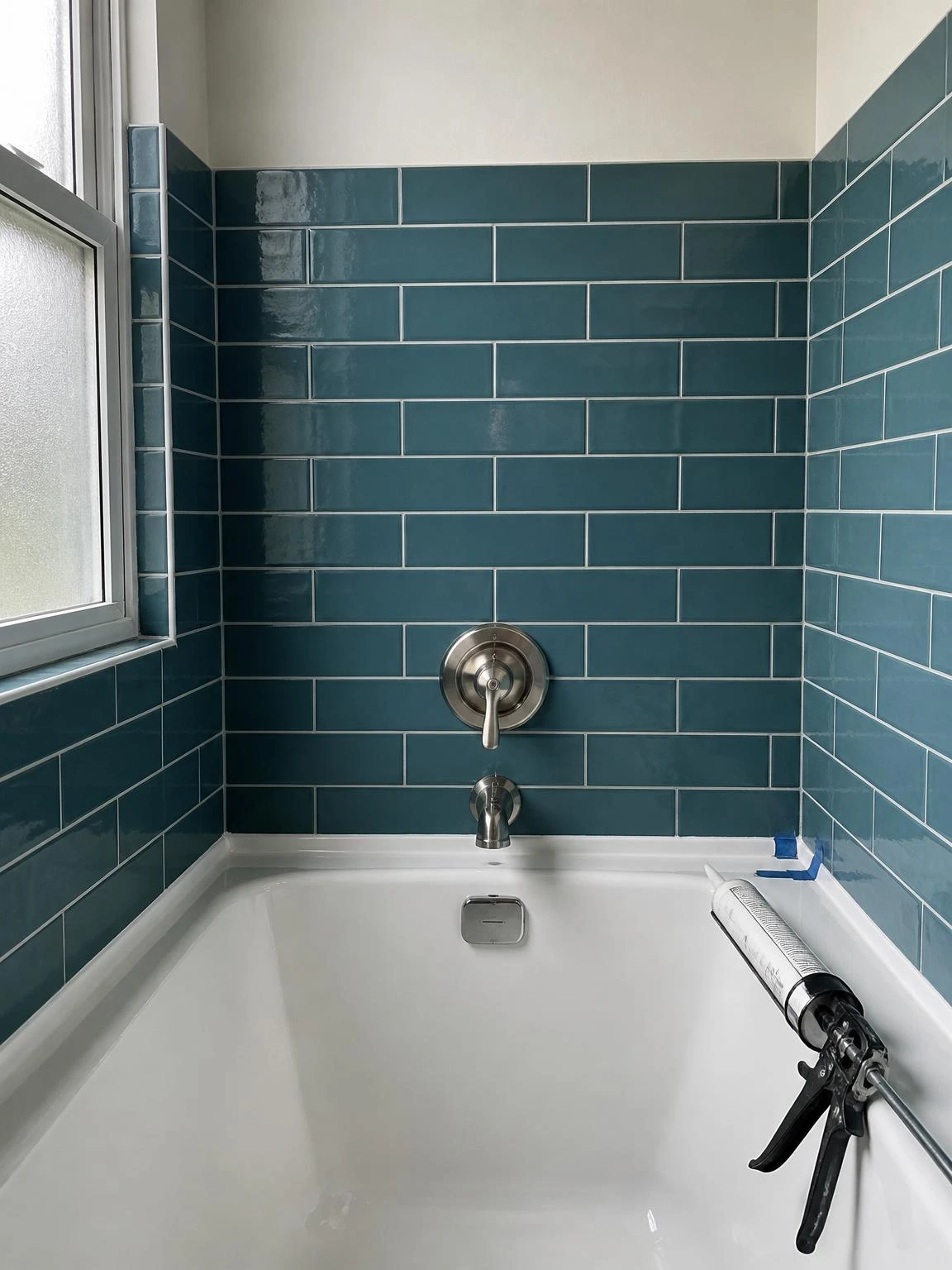Fresh white silicone caulk bead sealing the tub-to-tile joint in a teal subway tile bathtub surround, with painter's tape mid-removal