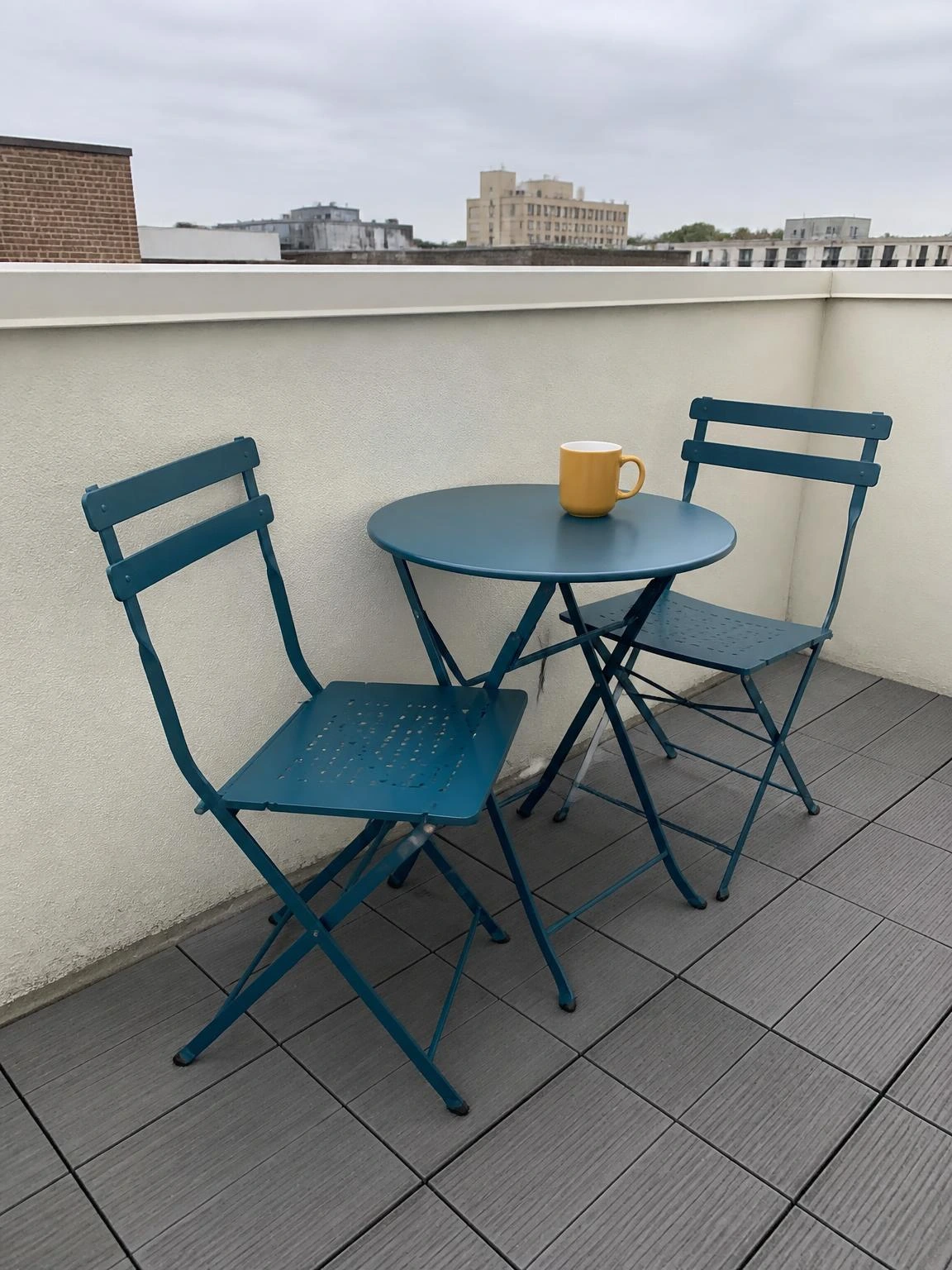 Deep teal bistro table and two folding chairs scaled for a compact rooftop terrace, with urban skyline visible above the white parapet wall