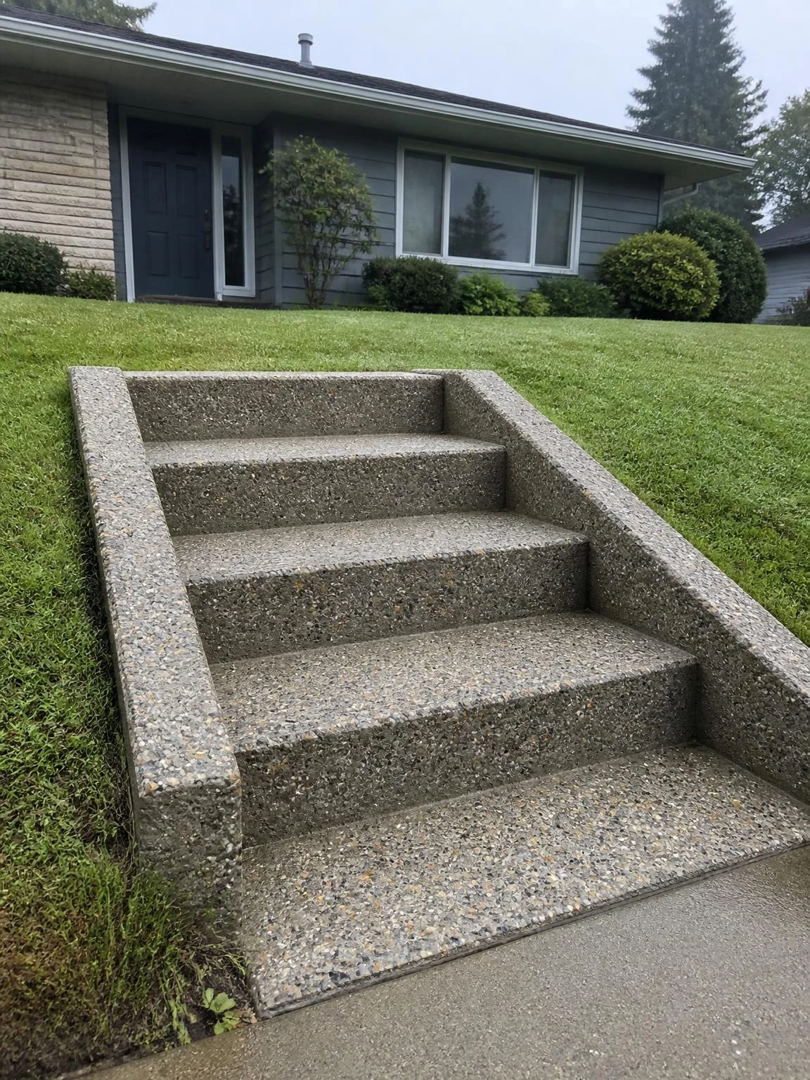 Four wide poured concrete steps cut into a sloped front yard, with exposed aggregate treads and matching flanking retaining walls that blend seamlessly with the home foundation