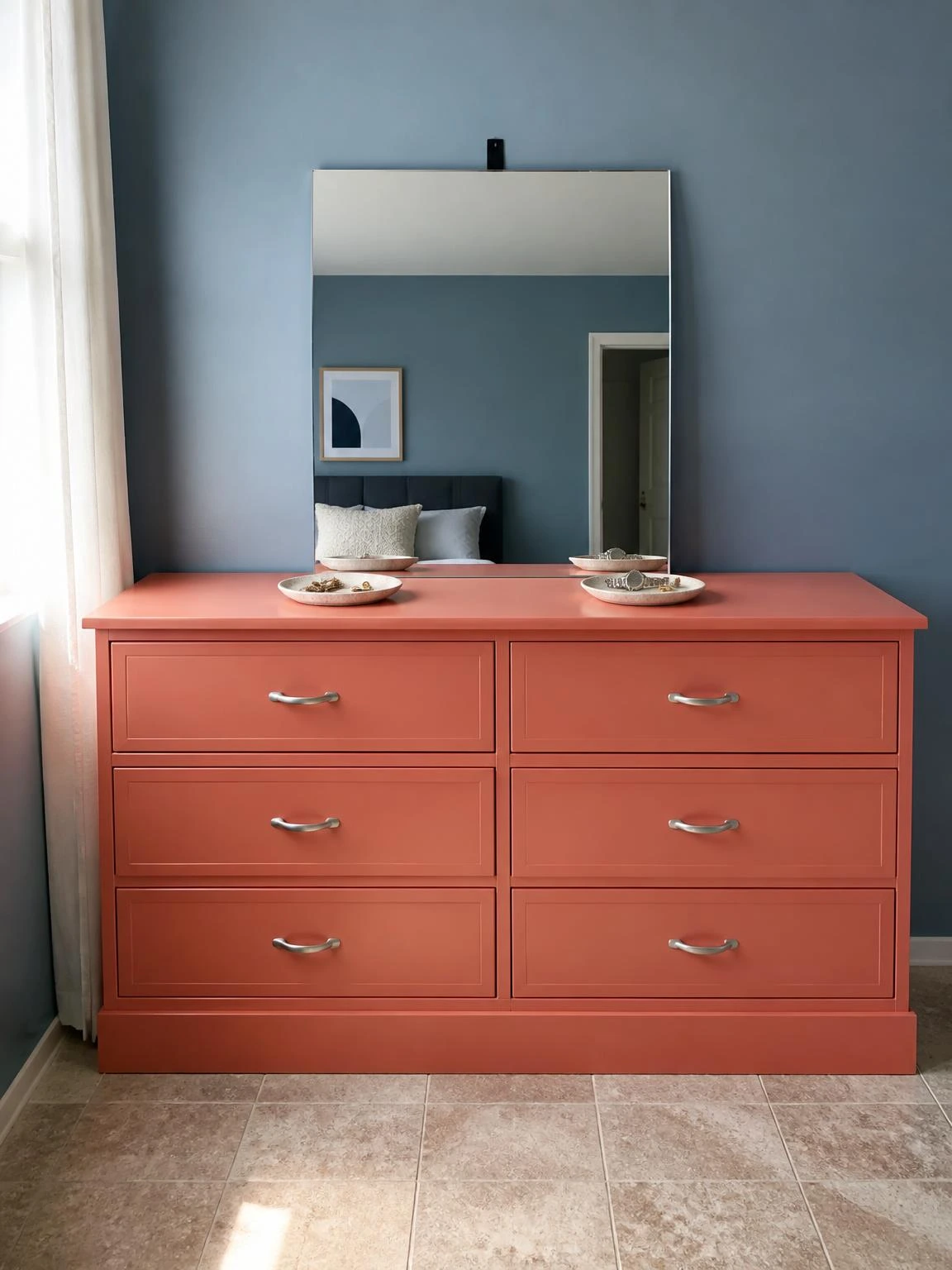 Soft coral six-drawer dresser against a muted blue wall styled with a leaning mirror and two small ceramic trays holding jewelry and a watch for couple room decor