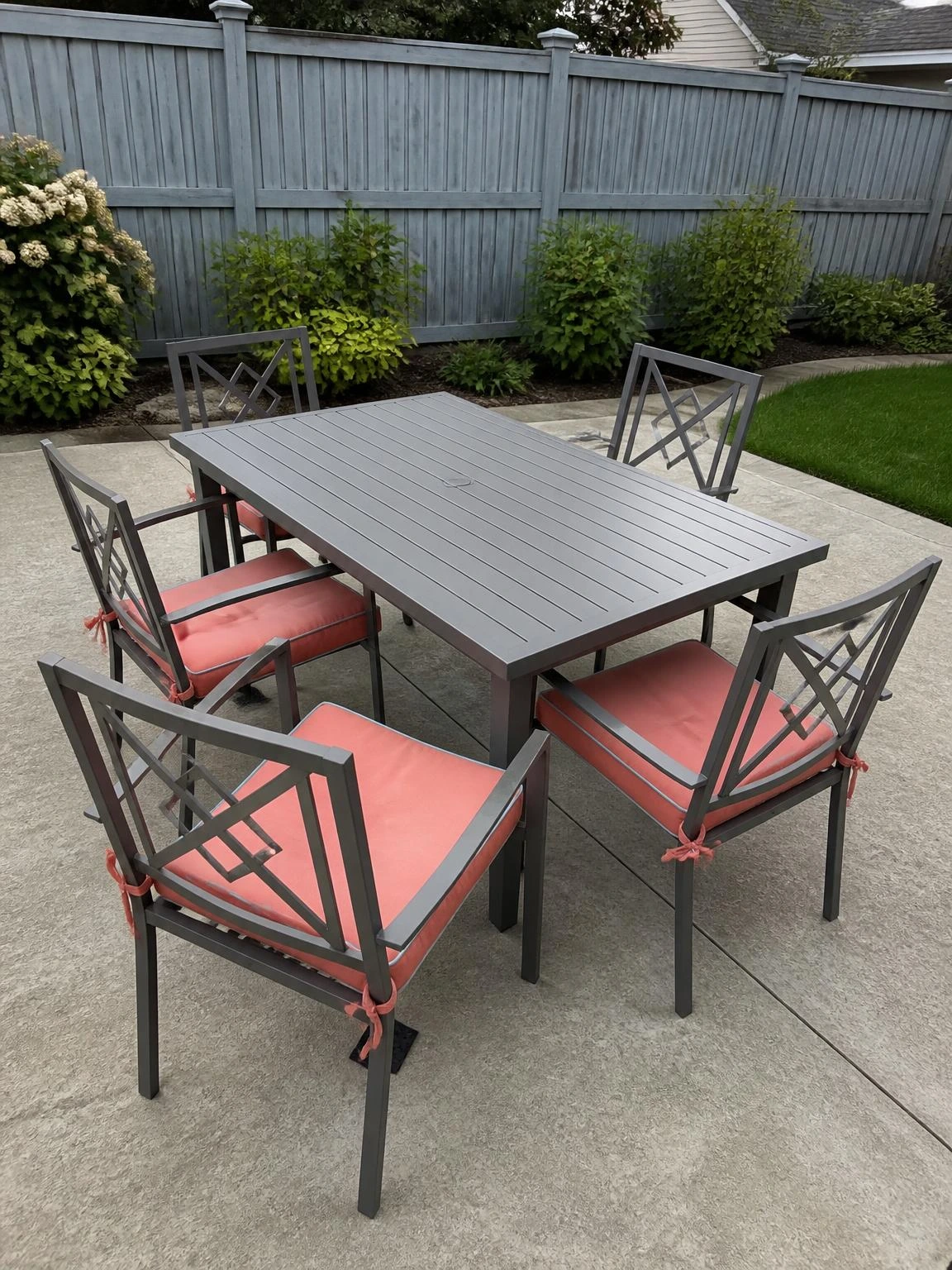 Concrete backyard patio with four metal dining chairs fitted with tied-on soft coral outdoor cushions around a rectangular table, a simple summer upgrade