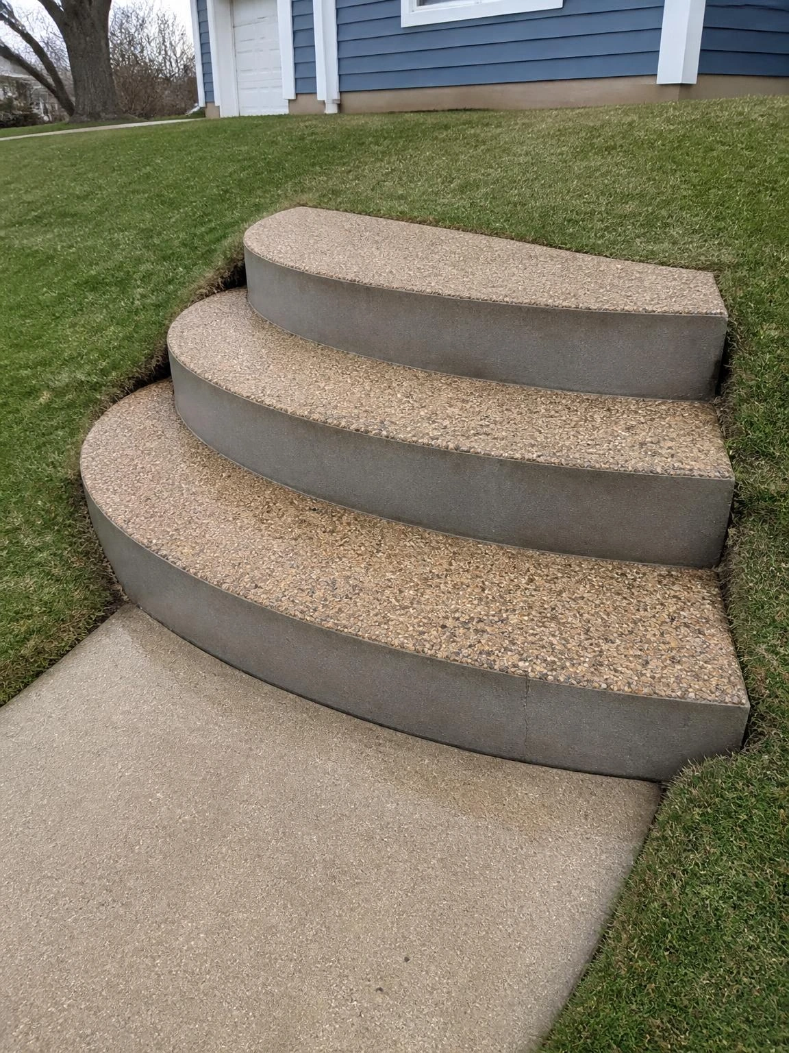 Curved concrete landscape stairs with exposed aggregate treads sweep across a rounded front yard slope, showing how landscape stairs can follow natural terrain geometry