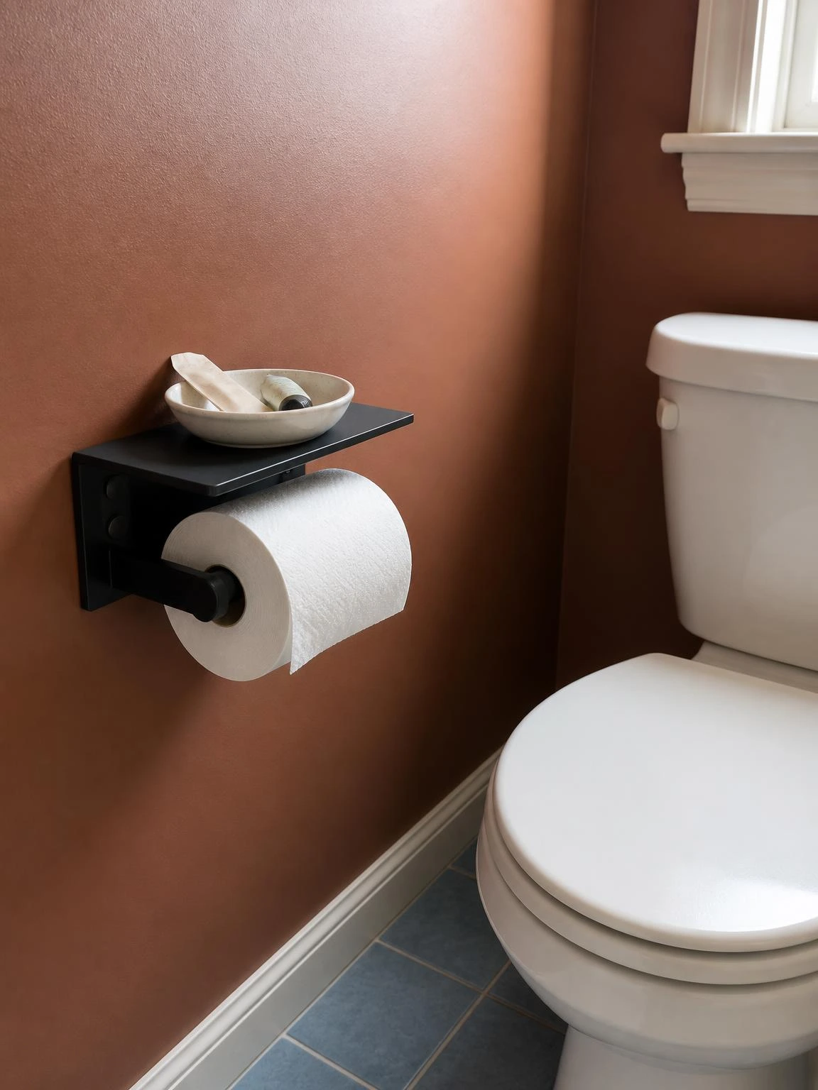 Matte black toilet paper holder with integrated shelf mounted on rust wall beside white toilet, a budget bathroom hardware upgrade that adds storage