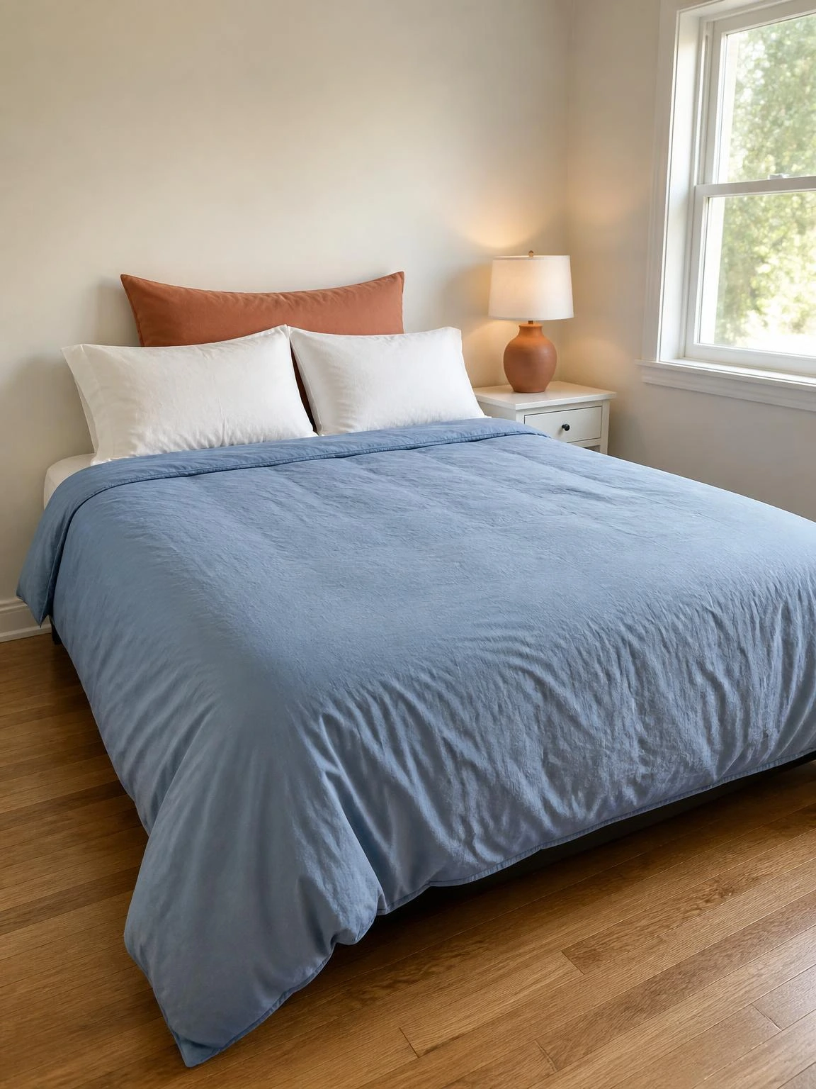 Queen bed dressed in a dusty blue cotton percale duvet with a terracotta euro sham, showing an easy summer bedroom refresh against warm white walls