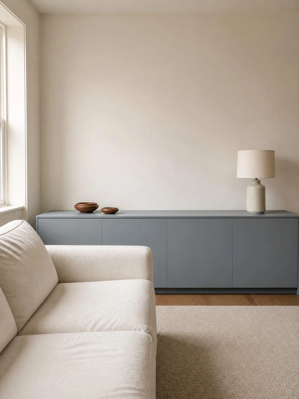 Low dusty blue credenza with flush hardware-free doors anchoring a minimal living room, styled with a ceramic lamp and terracotta dishes