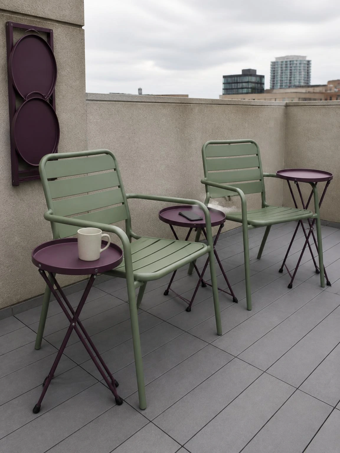 Three muted plum folding tray tables beside green metal chairs on a grey composite rooftop deck, showing fold joints and wall-mounted storage rack for small rooftop terrace design
