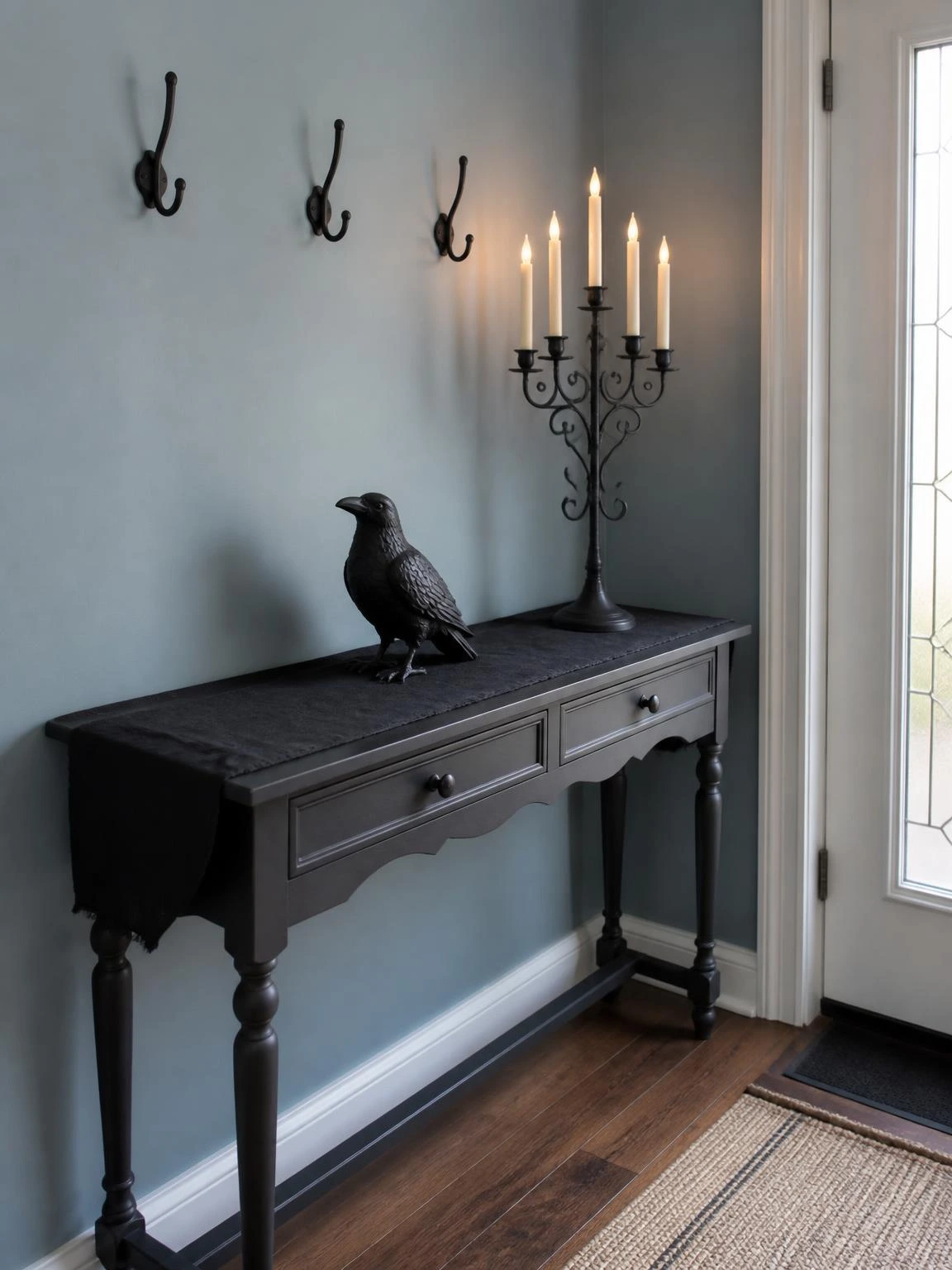 Gothic entryway console table styled with a black iron candelabra, ceramic raven figure, and black cotton runner against a dusty blue wall