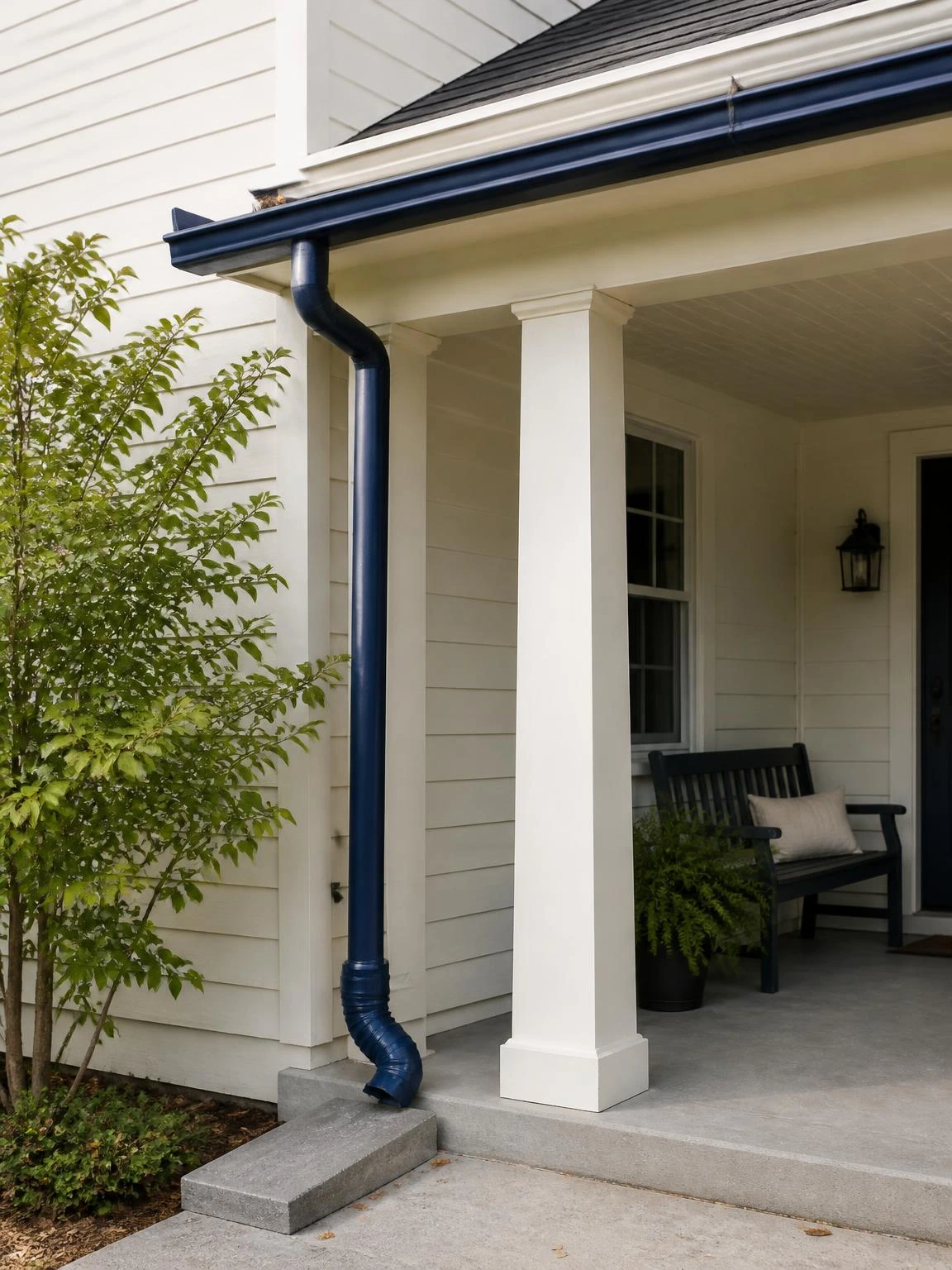 Navy K-style aluminum gutter and matching downspout integrated at the front corner of an ivory porch roof, directing runoff safely away from the column base