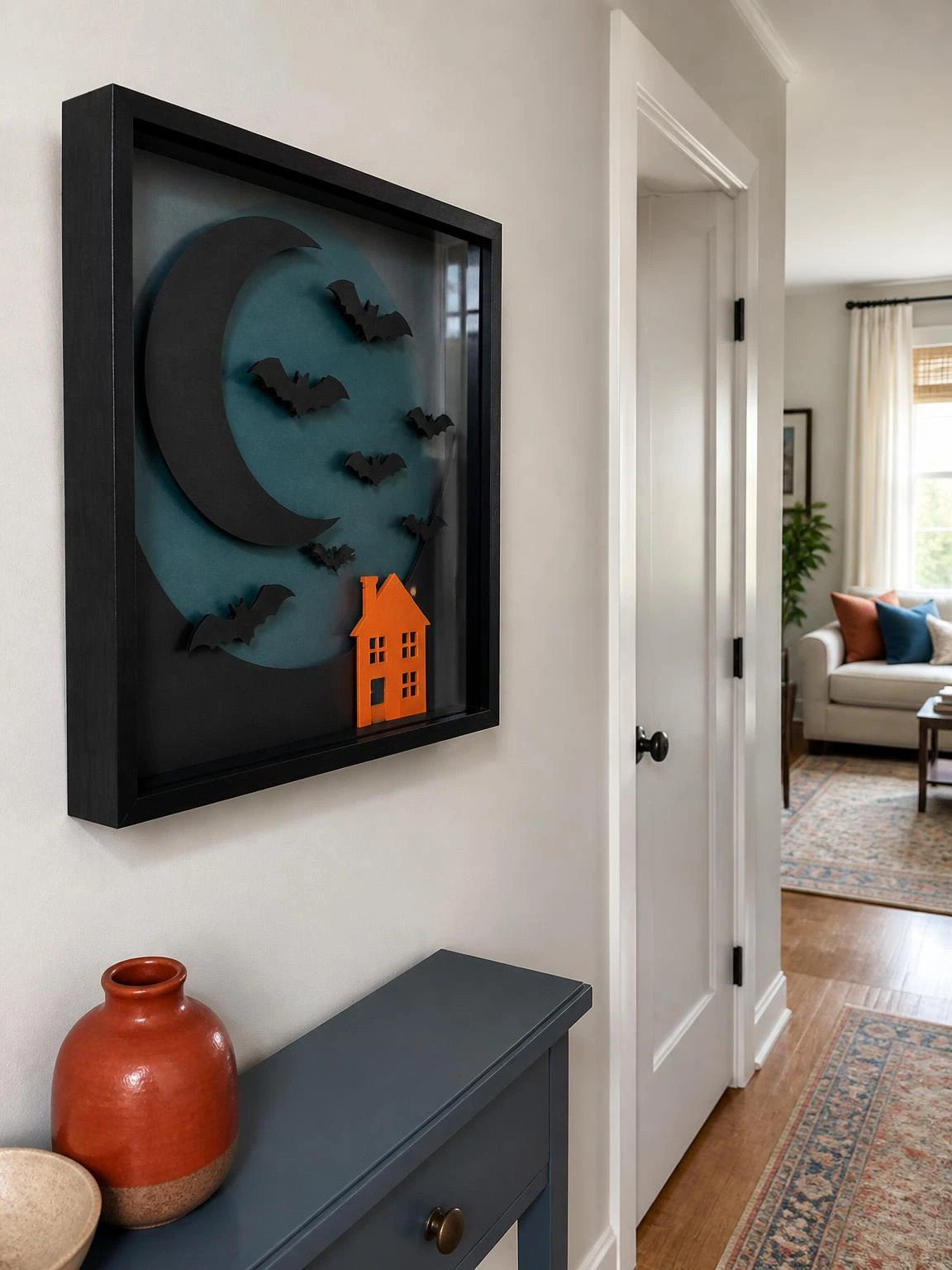 Halloween crafts DIY project: layered teal moons, black bats, and an orange house fill a framed shadow box in a bright hallway.