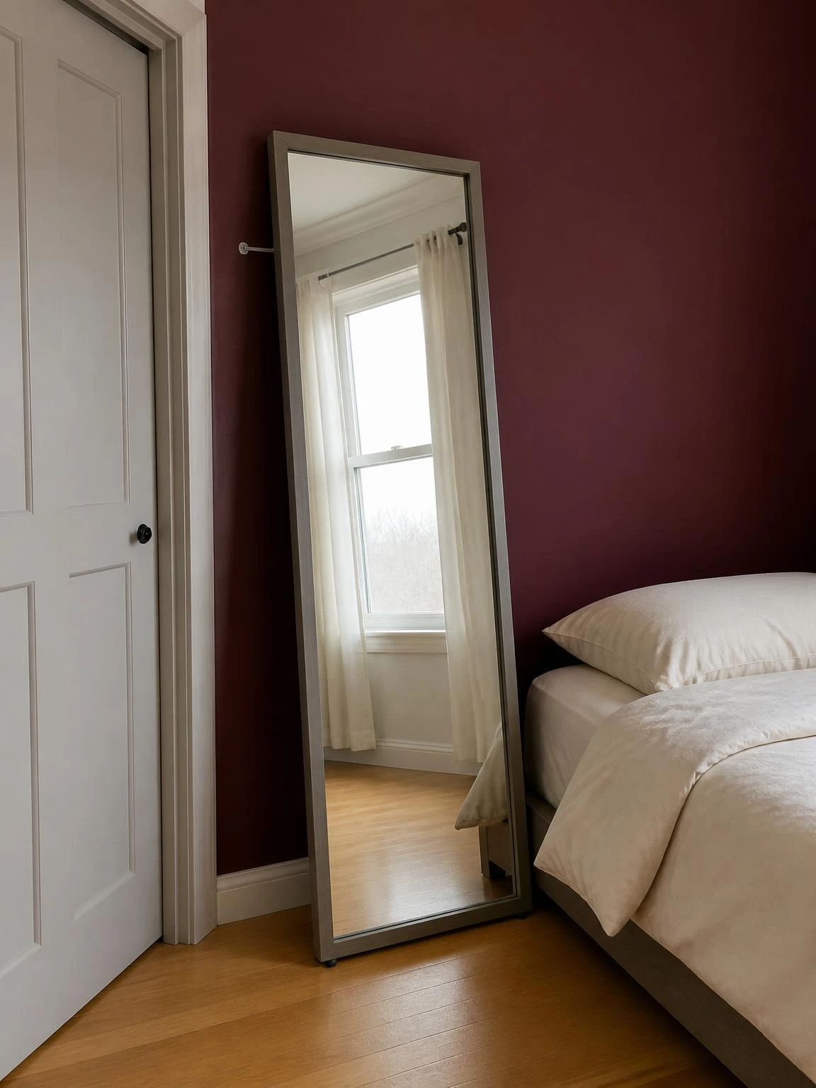 Full-length mirror with slim gray frame leaning against a burgundy bedroom wall, reflecting window light to visually expand a compact space