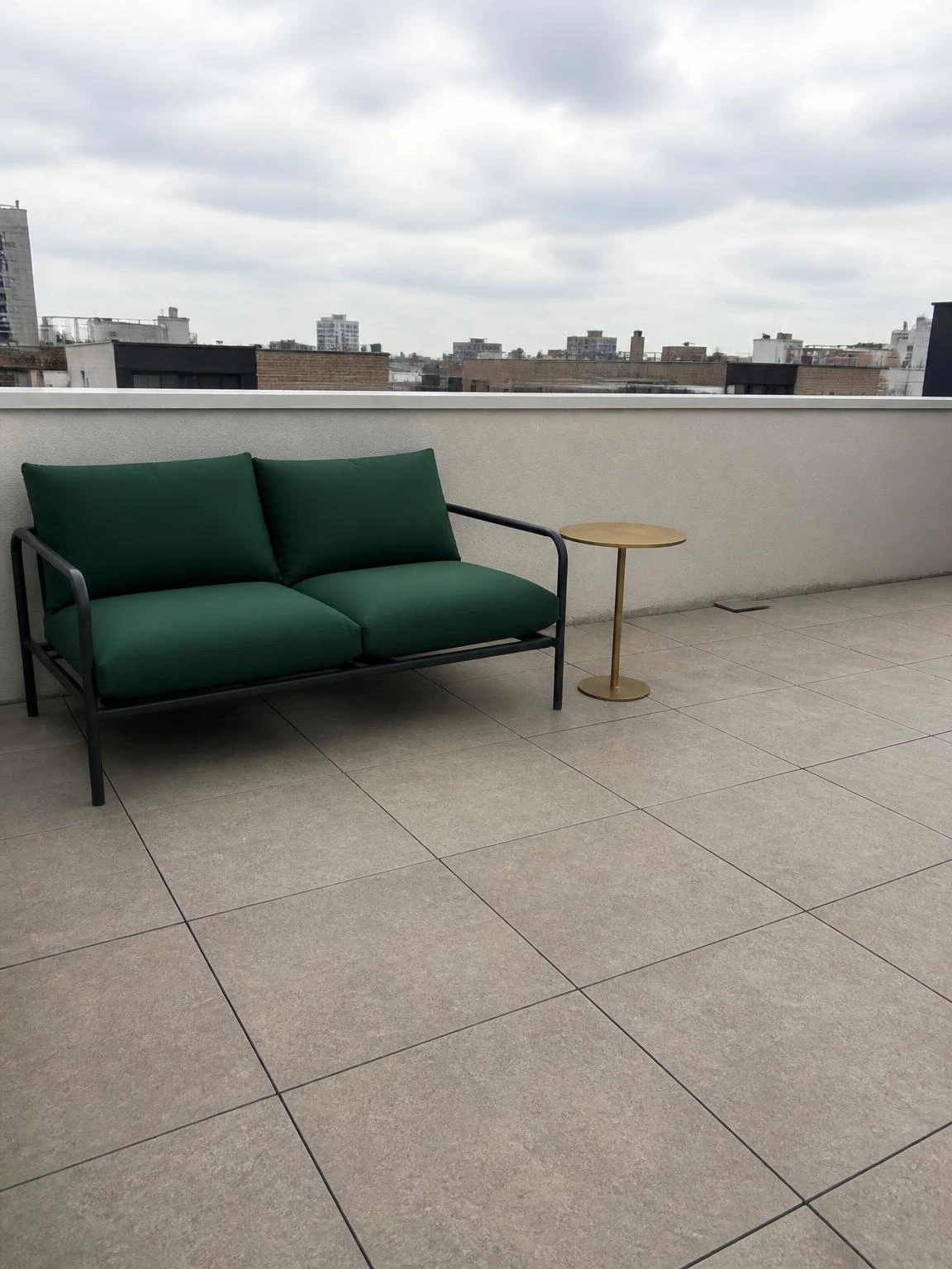 Snap-together porcelain deck tiles in warm stone-grey cover a compact urban rooftop terrace floor, paired with a forest-green loveseat and brass side table