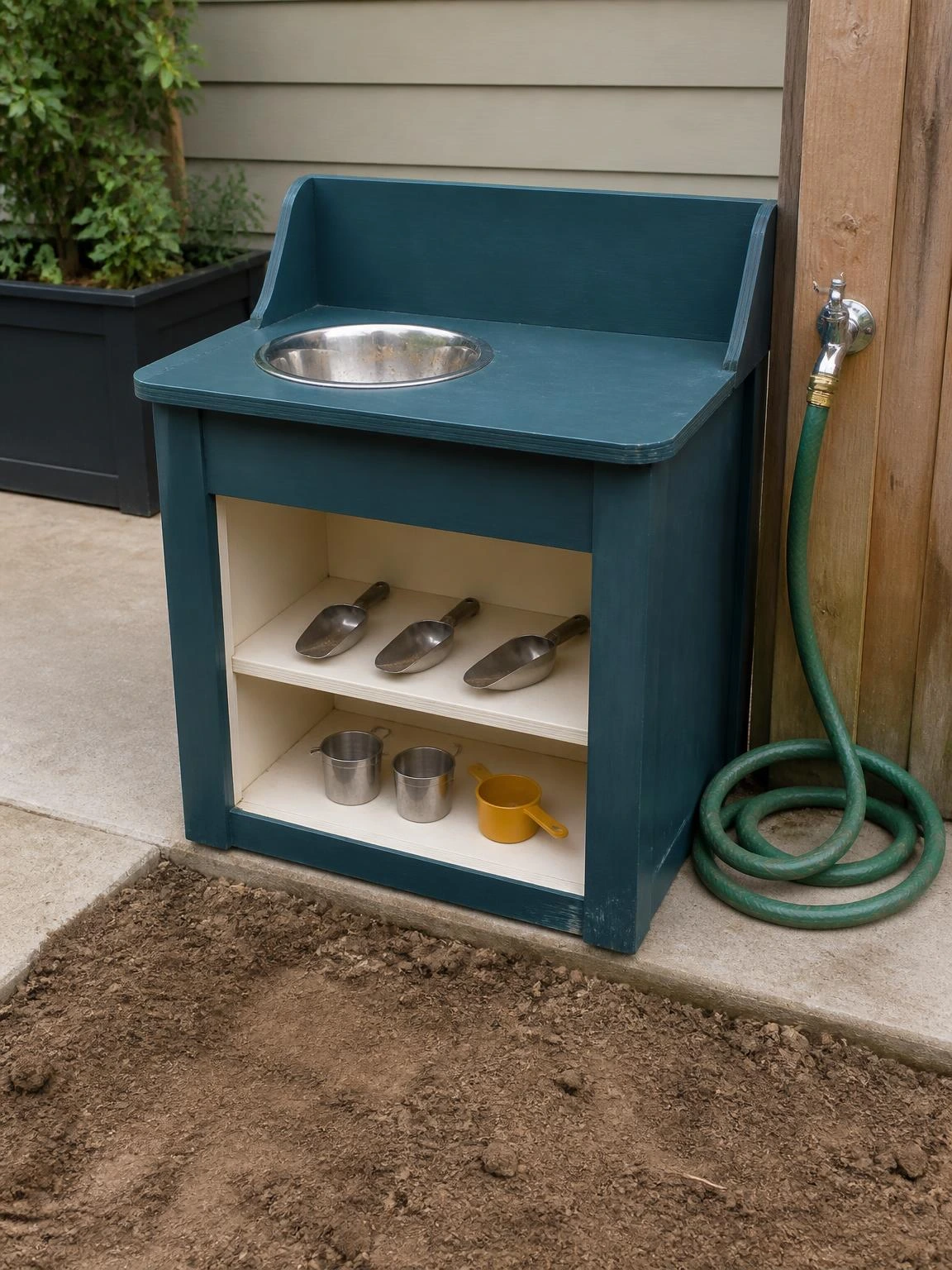 Teal plywood mud kitchen station with built-in stainless bowl and open shelving holds metal scoops at a concrete patio edge, ready for kids' outdoor sensory play