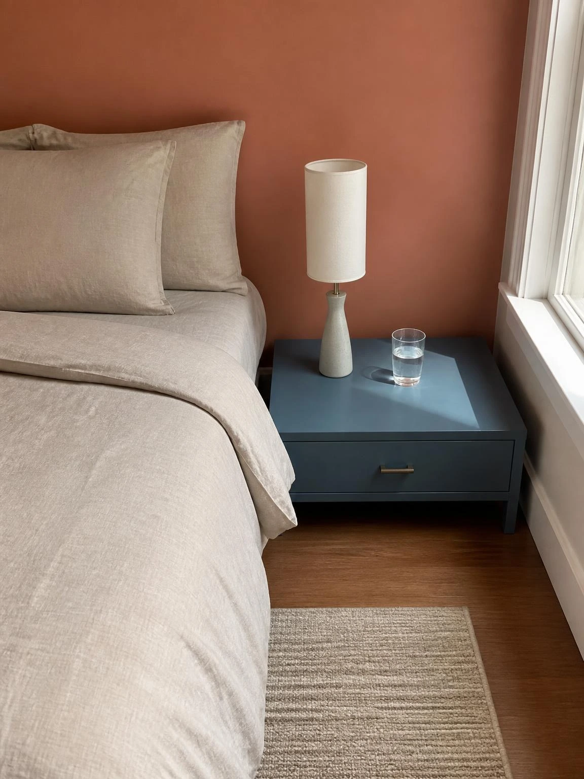 Muted blue bedside table against a coral wall holding only a ceramic lamp and glass of water — a minimal home decor setup done right