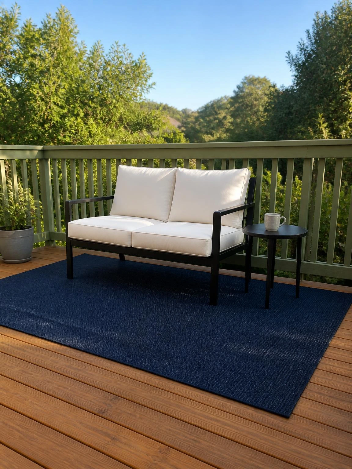 Navy flat-weave outdoor rug anchoring a black metal loveseat on a honey-toned timber deck, styled for a relaxed summer deck setup
