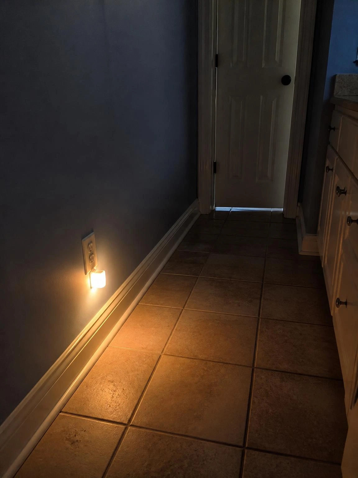 Plug-in LED night light glowing in a low bathroom hallway outlet, casting soft amber light onto floor tiles without disturbing sleep
