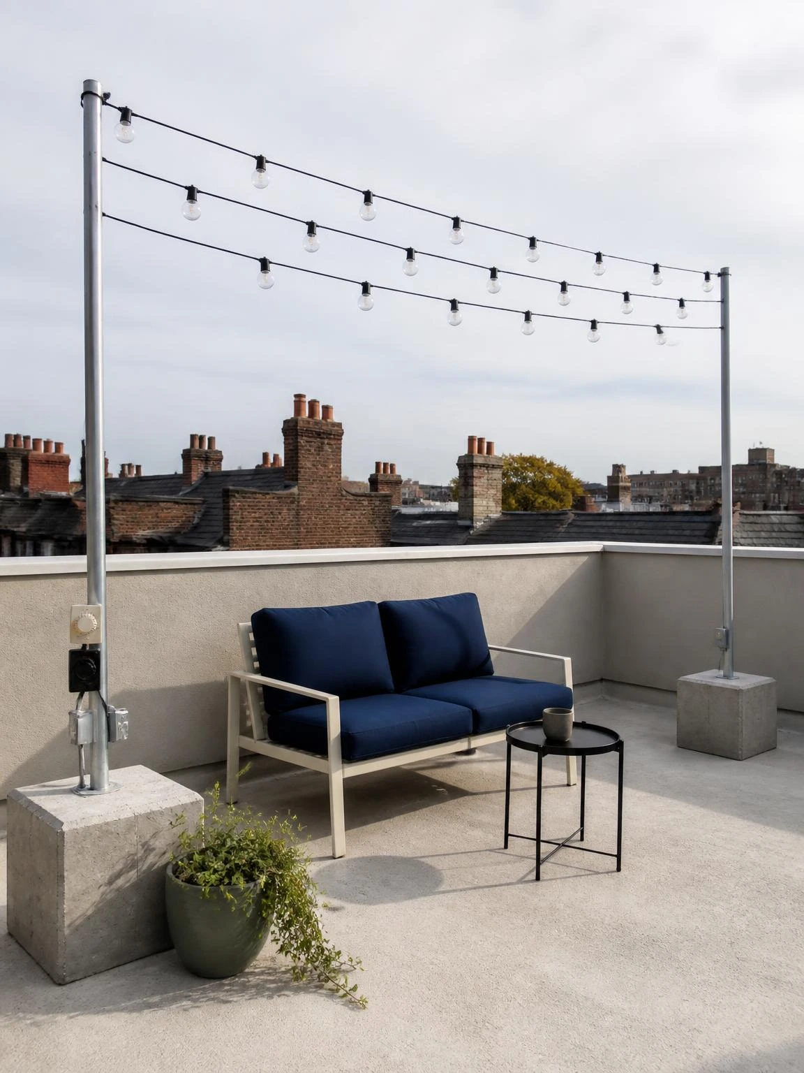 Overhead string light canopy on a small rooftop terrace, clear glass bulbs strung between two galvanised poles in concrete ballast bases above a navy-cushioned sofa