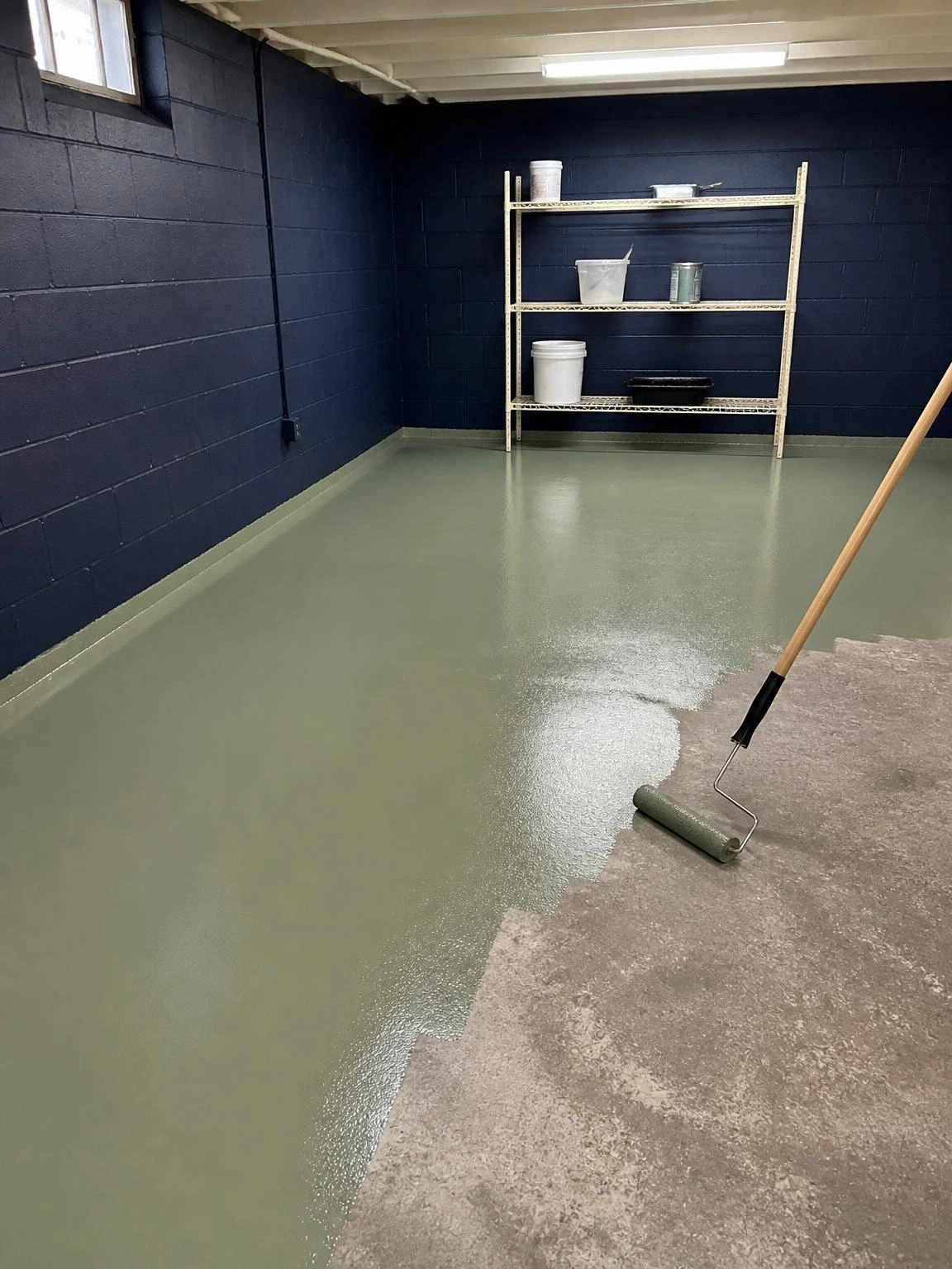 Basement concrete floor mid-application with muted sage-green epoxy paint showing a sharp wet edge where rolled coverage meets bare gray concrete