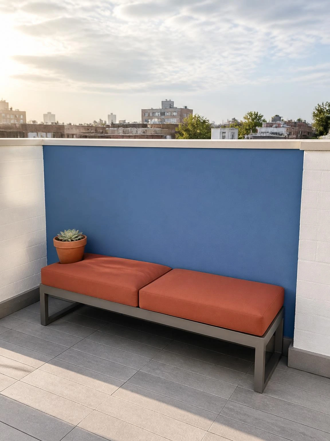 Compact rooftop terrace with a dusty blue painted parapet accent wall backdrop, low aluminium bench with terracotta cushions, and a single succulent pot on grey porcelain tile