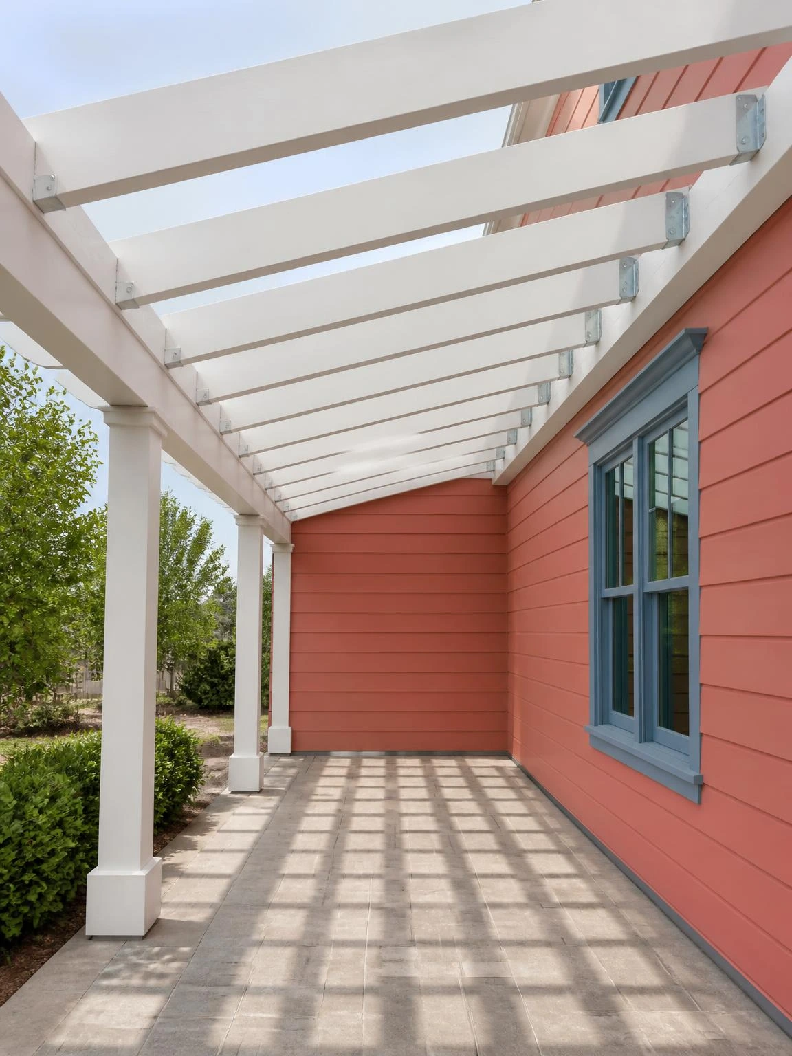 Pergola-style open porch roof with evenly spaced white painted rafters seated in galvanized joist hangers, casting parallel shadow bars across stone-look pavers below