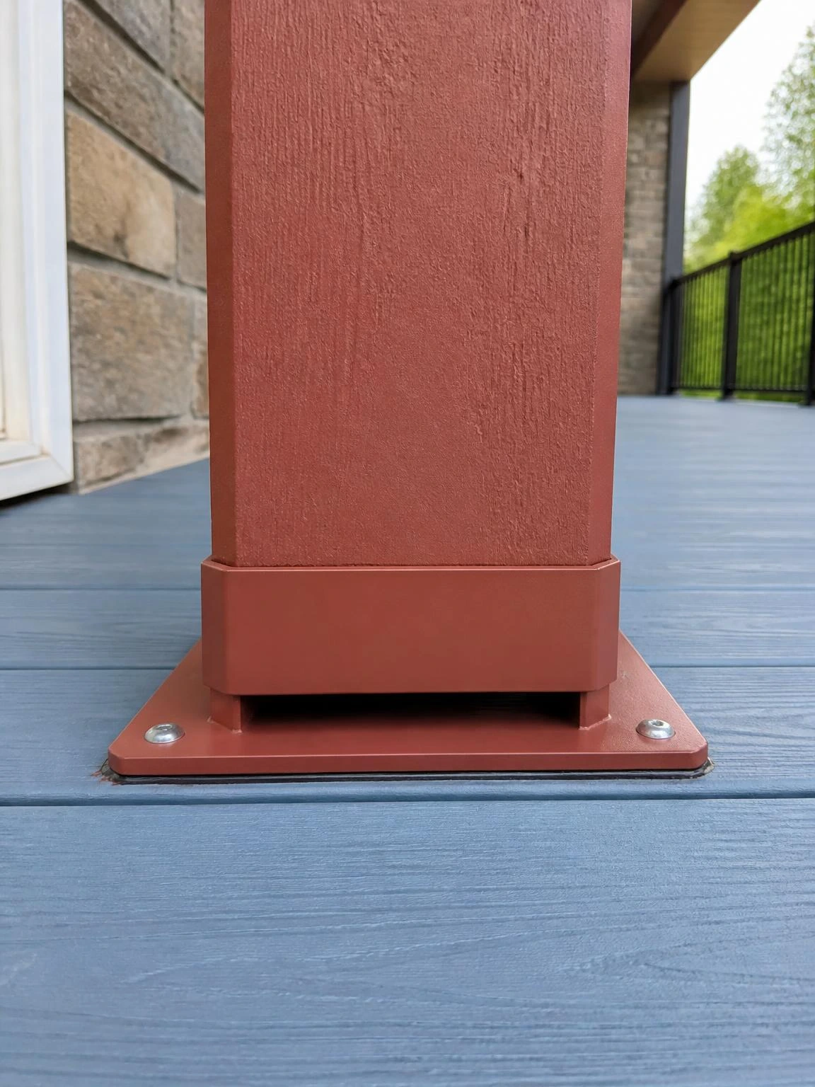 Powder-coated aluminum standoff base connector elevates a rust-red porch column above blue composite decking, with butyl tape flashing sealing the plate for moisture drainage protection