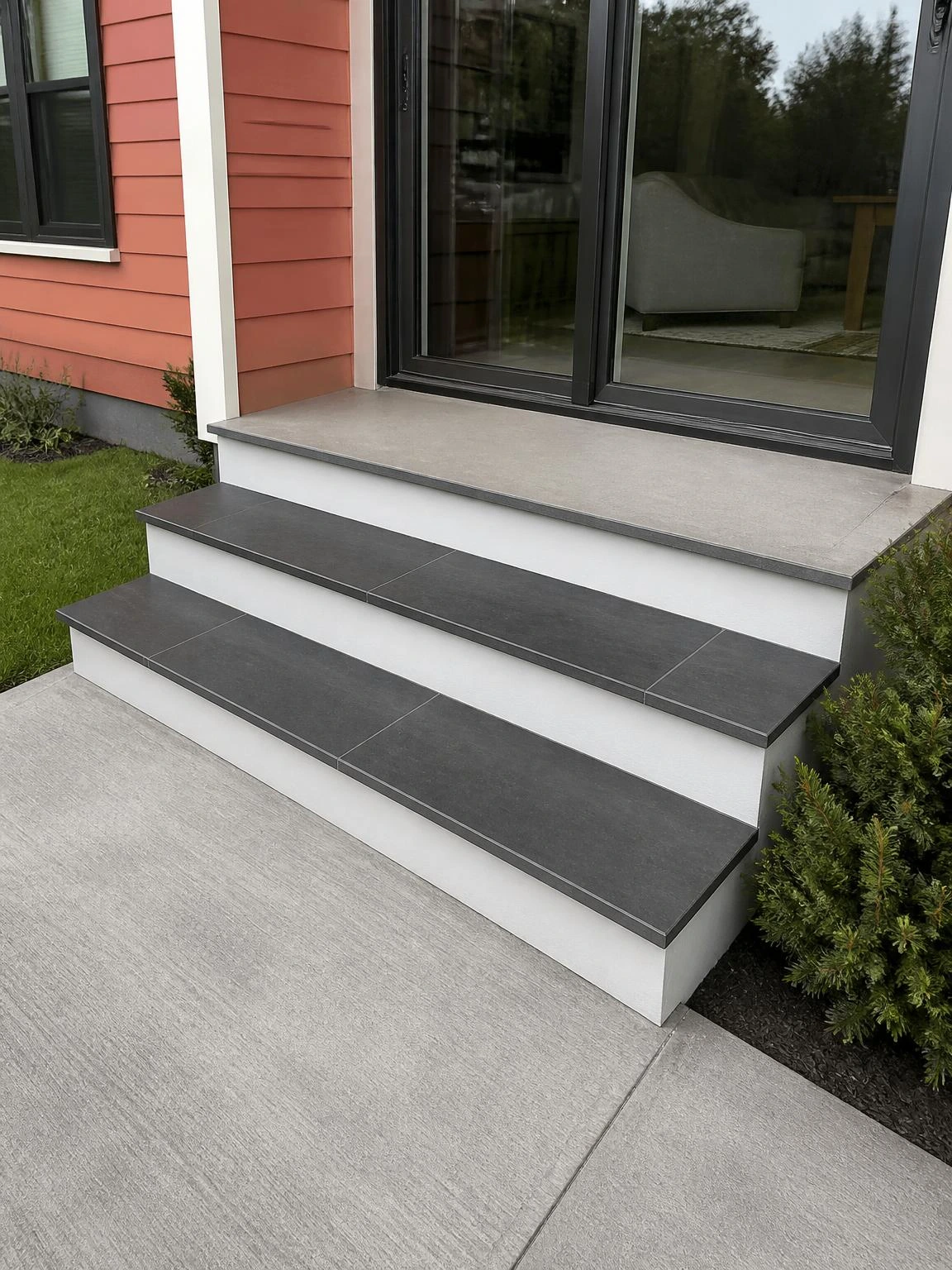 Three wide poured concrete landscape stairs with charcoal porcelain tile tread inlays leading to a rear patio sliding glass door, shown in soft afternoon light