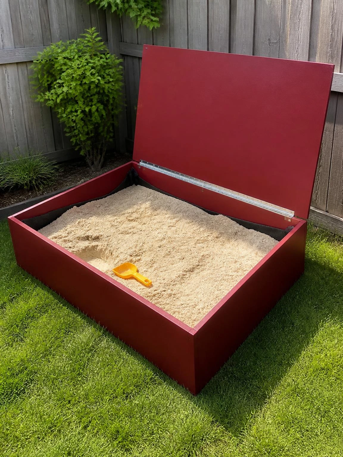 Burgundy-painted raised sandbox with hinged lid propped open in a sunlit backyard corner, showing sand depth and drainage fabric inside