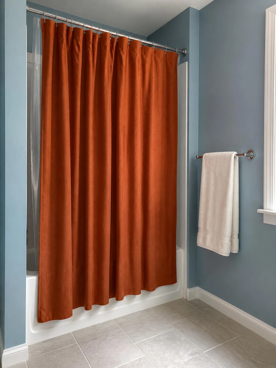 Rust-orange cotton shower curtain hanging from a chrome rod over a tub-shower combo, adding bold color to soft blue bathroom walls on a budget