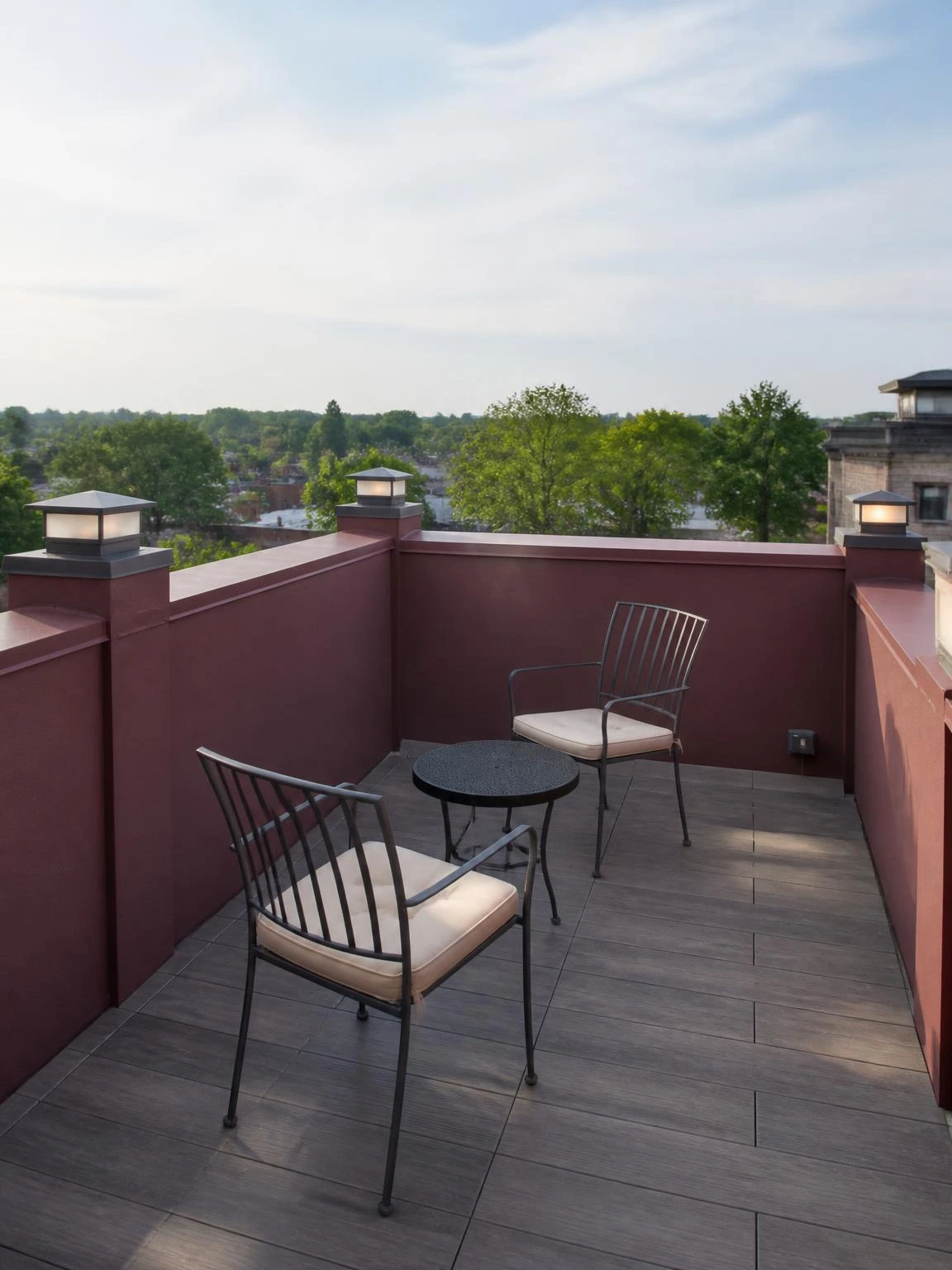 Four matte gunmetal solar post cap lights mounted on burgundy plaster parapet posts of a compact rooftop terrace, emitting a soft glow in late-morning daylight