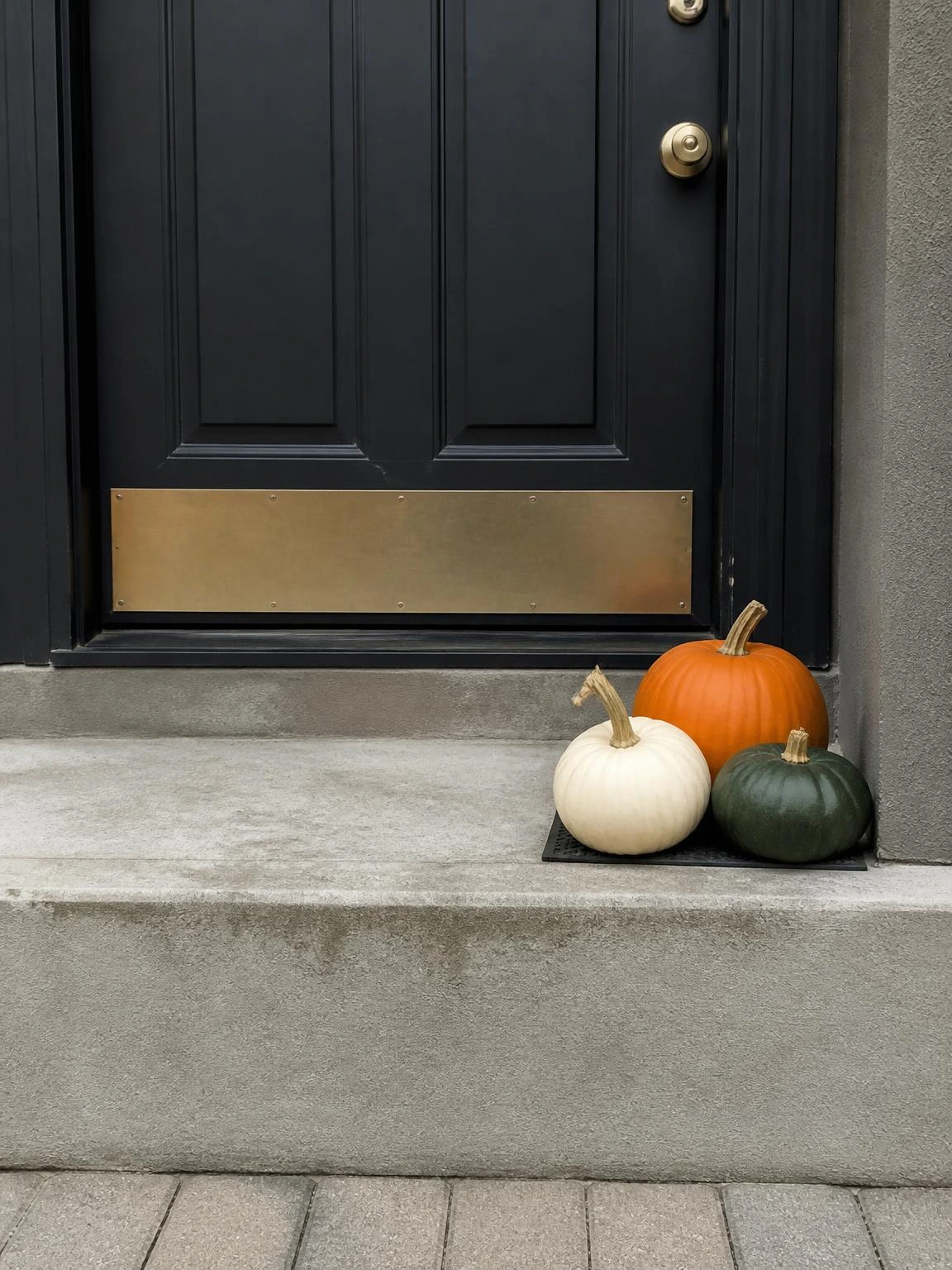 Three mini pumpkins in burnt orange, white, and forest green clustered on a narrow concrete step against a charcoal door — a small porch halloween idea that works in tight spaces