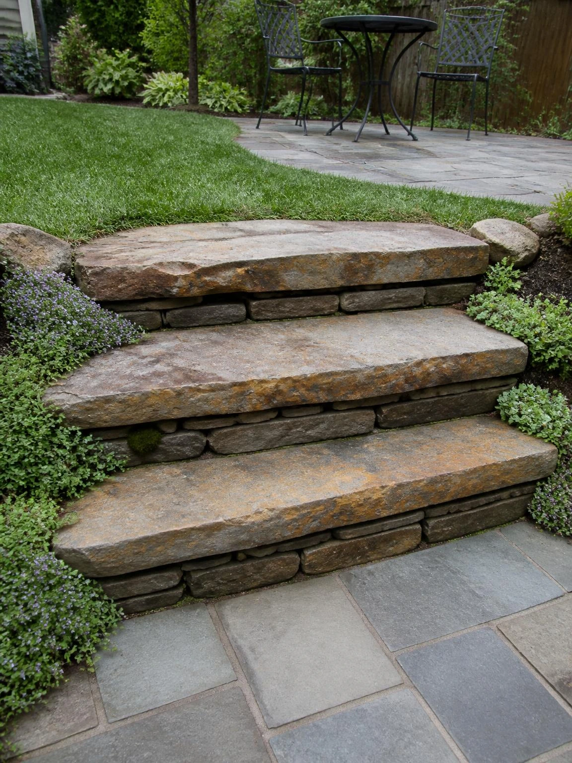 Dry-stacked fieldstone landscape stairs with rust-brown and gray treads descending to a bluestone patio, softened by creeping thyme and sedum along the risers