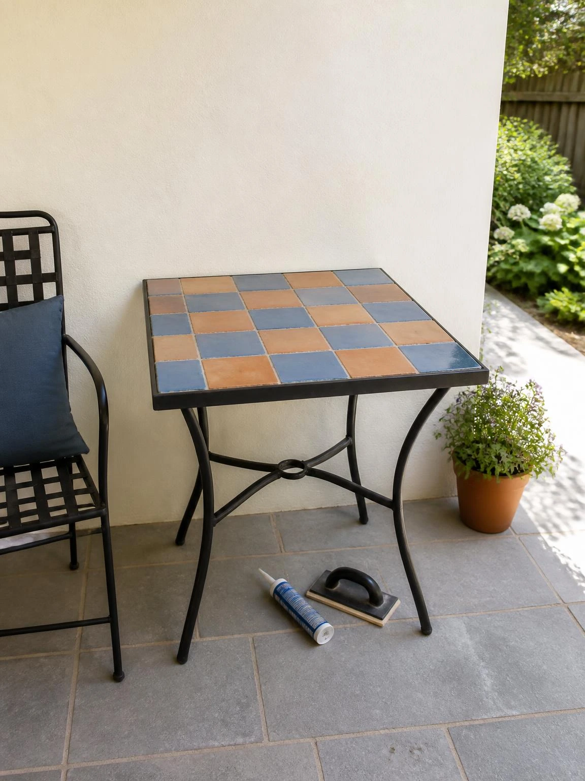 Outdoor tiled garden table with alternating terracotta and dusty-blue tiles beside a warm-white patio wall
