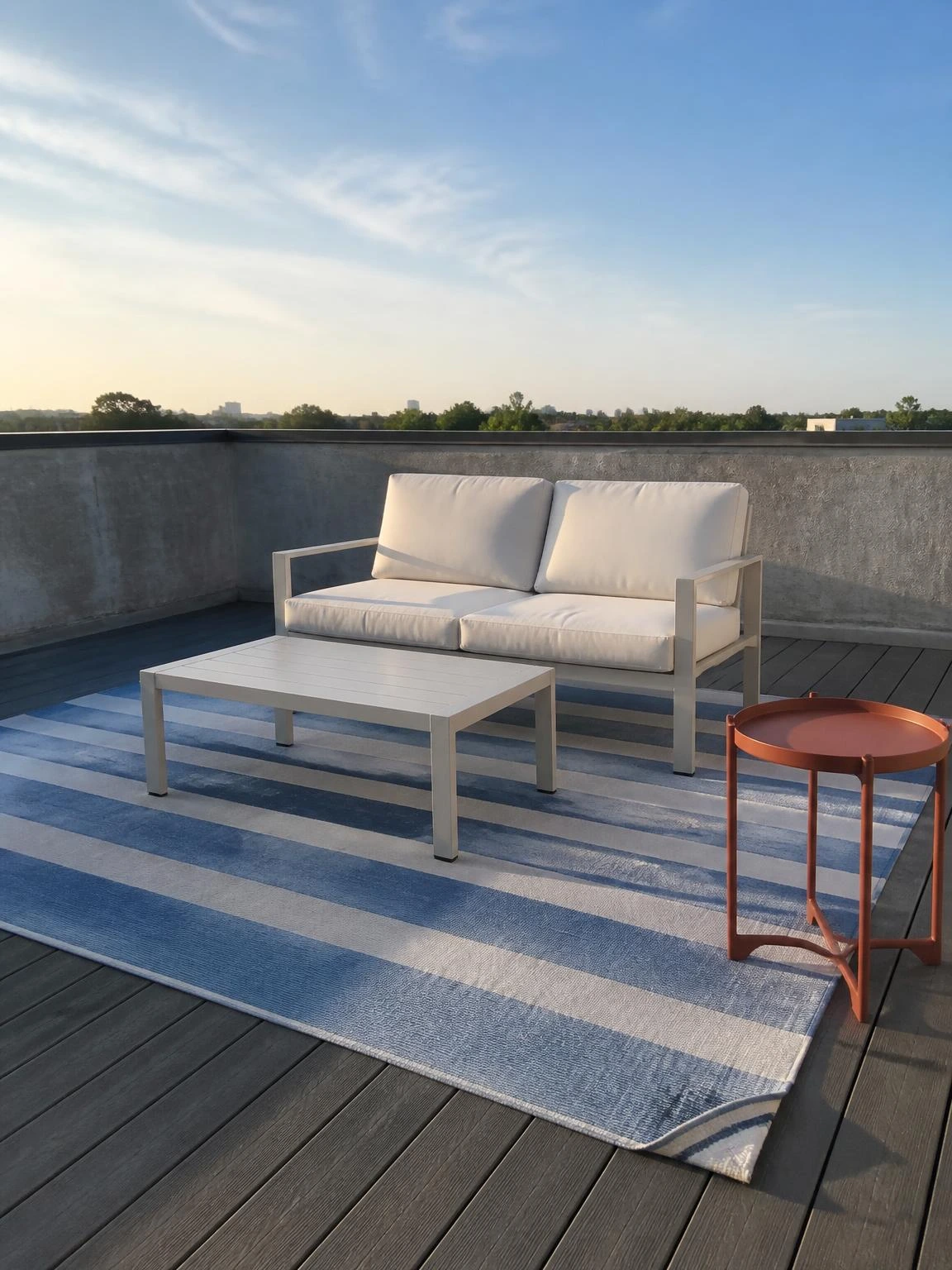 Dusty blue striped polypropylene outdoor rug anchoring a compact rooftop terrace seating zone on grey composite deck tiles in soft morning light