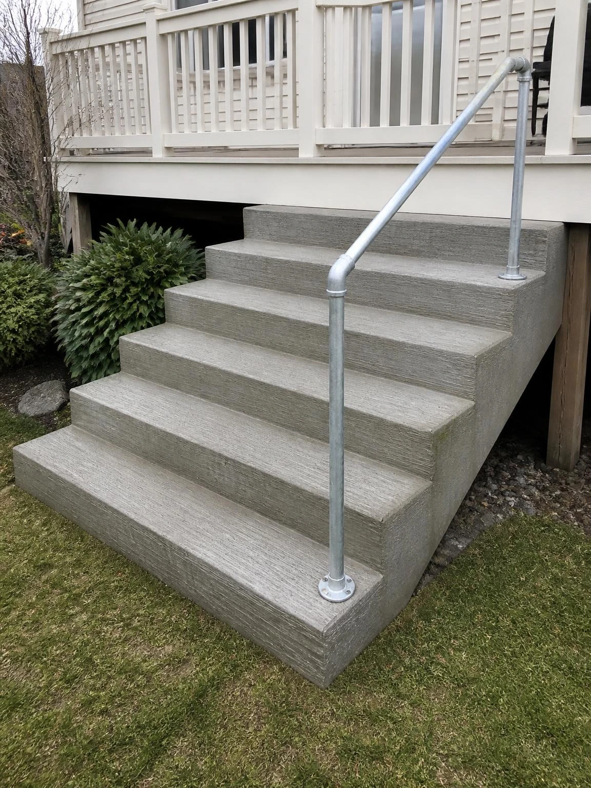 Six wide broom-finish concrete steps descend from a rear deck to a backyard, with a galvanized steel pipe handrail anchored along the right side