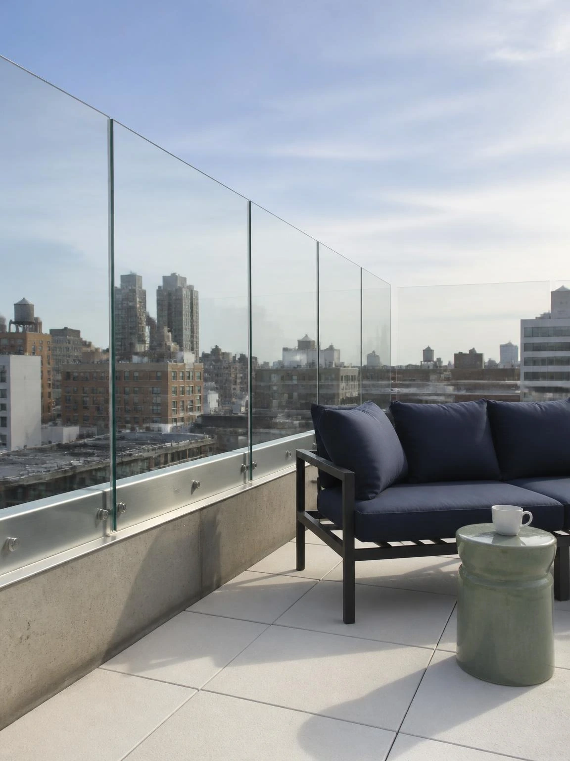 Frameless tempered glass windbreak panels in brushed stainless steel channels along a compact rooftop terrace parapet, city skyline visible through the clear low-iron glass